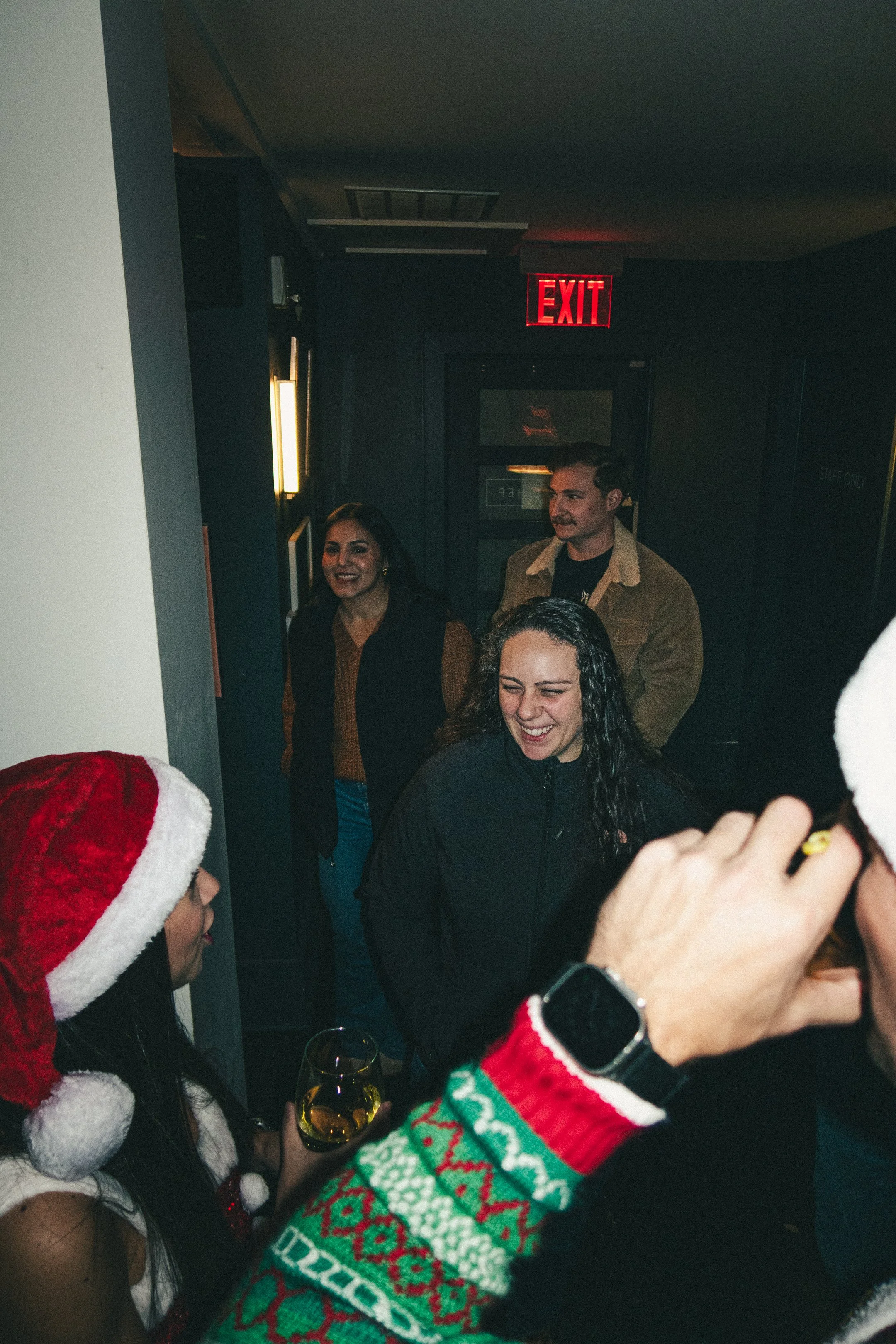 Group of people celebrating Christmas in a dimly lit room, some wearing Santa hats and festive sweaters, smiling and holding glasses of wine.