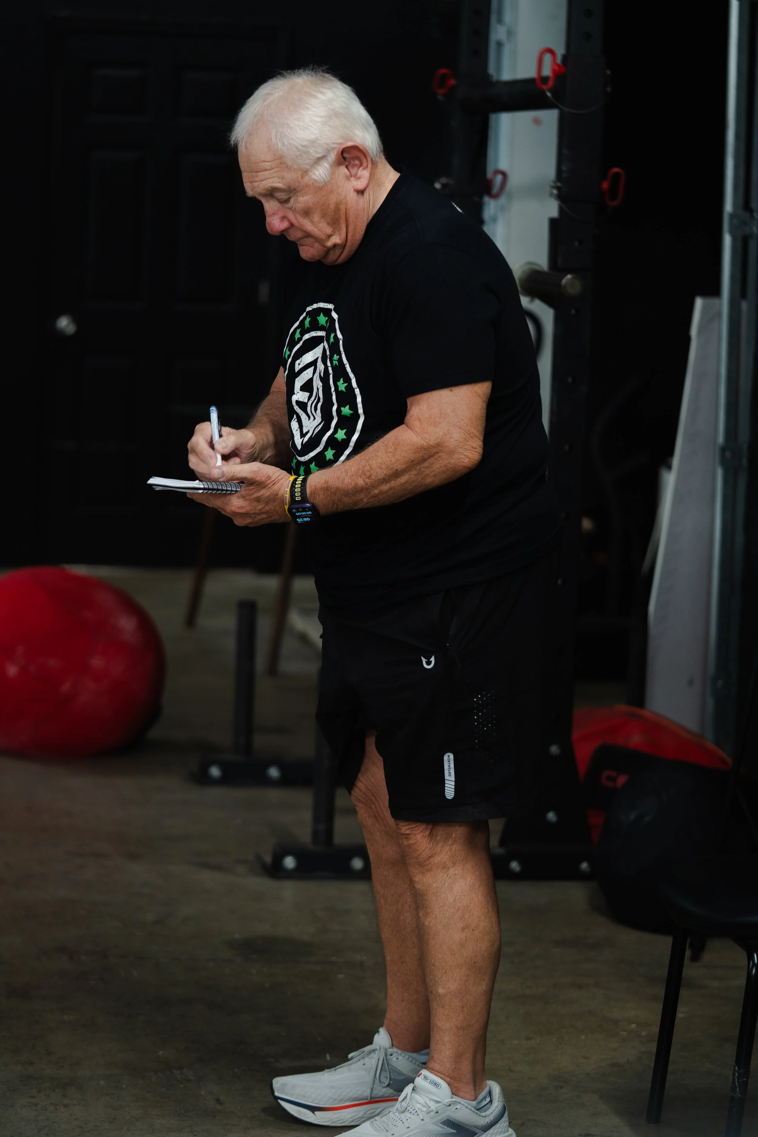 An elderly man with white hair is in a gym, writing in a notepad. He is wearing a black T-shirt with a logo, black shorts, white athletic shoes, and a smartwatch. The background includes gym equipment like medicine balls and weights.