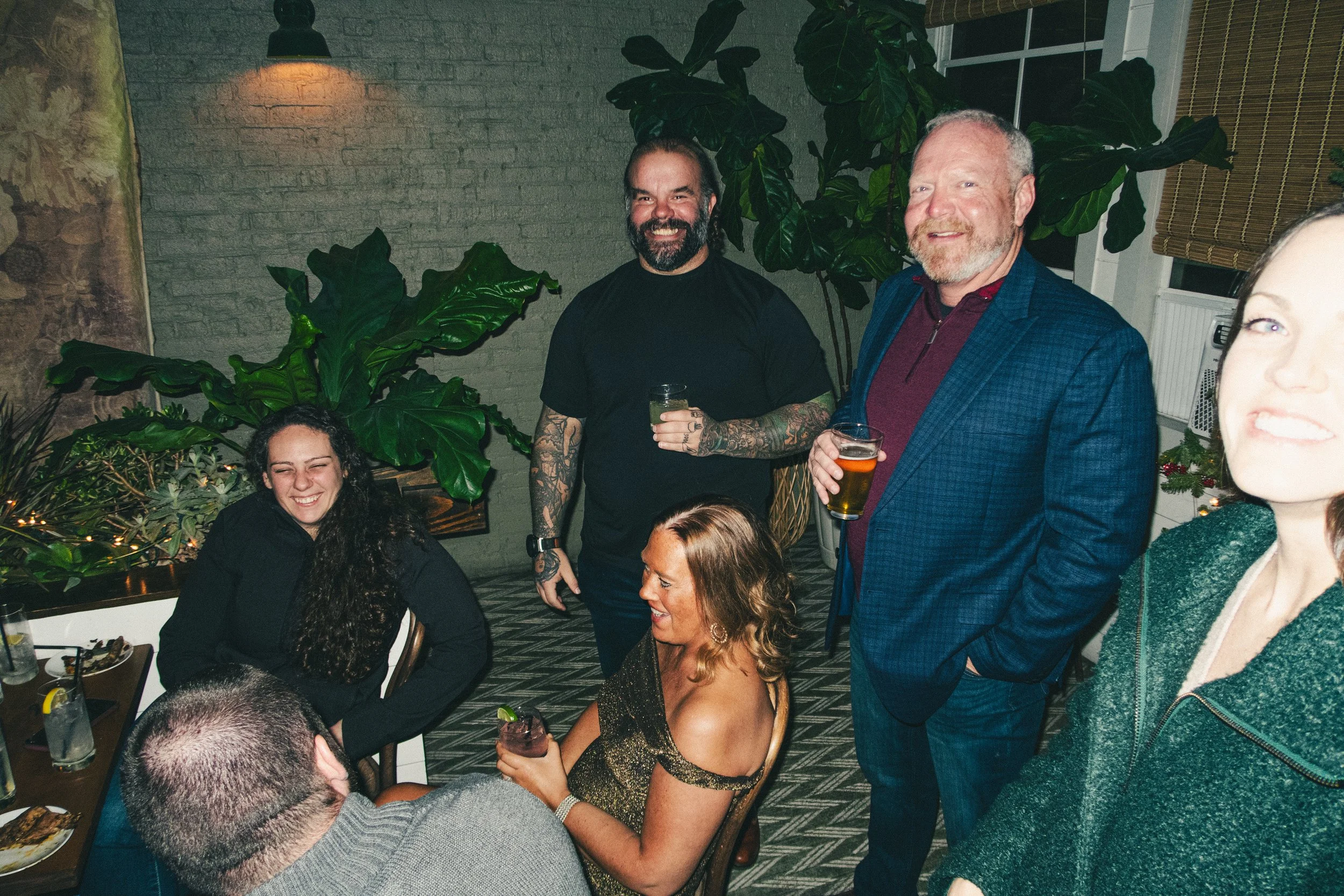 A group of people enjoying a party indoors with plants and artwork in the background, holding drinks and smiling.