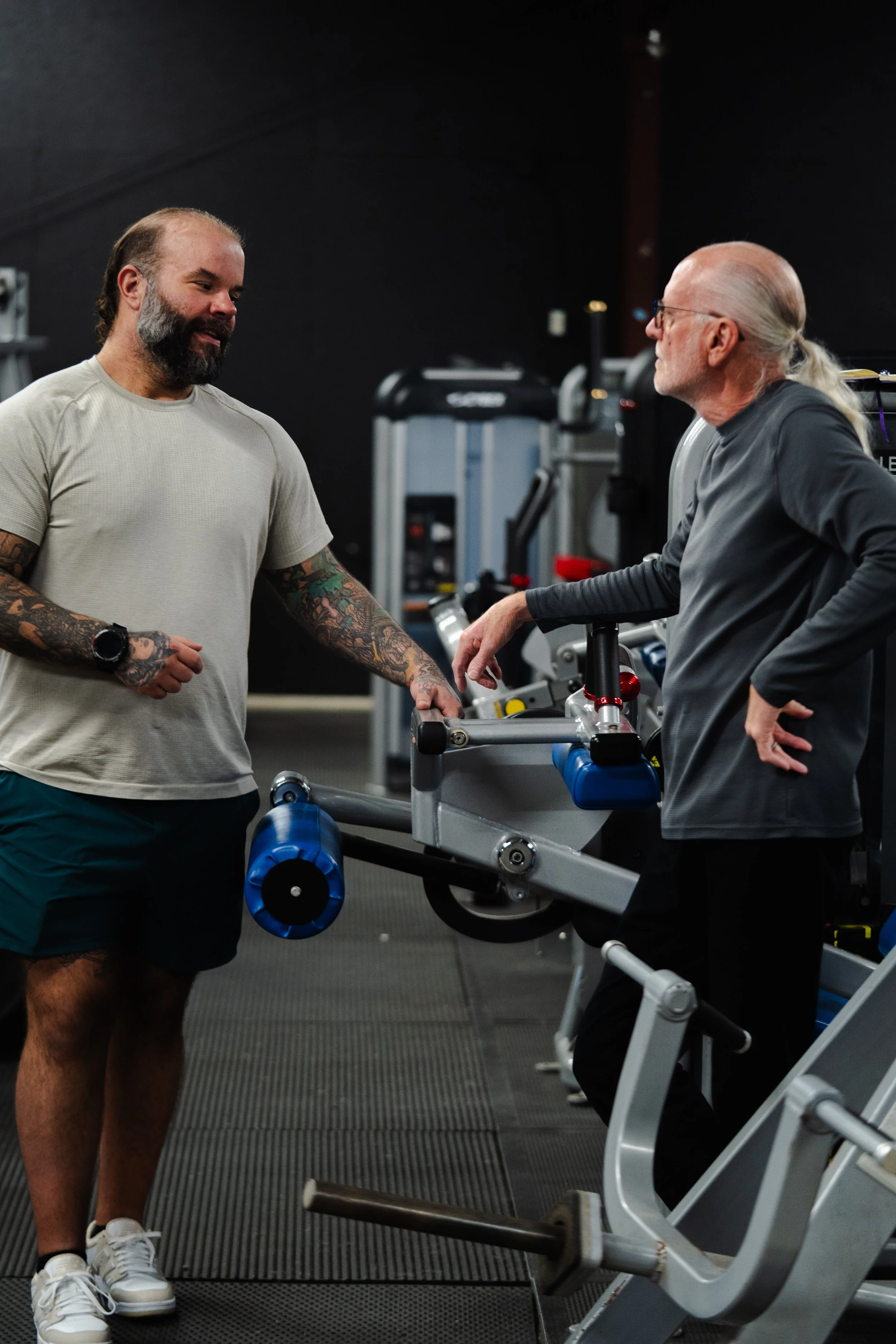 Two men in a gym, one with tattoos and a beard wearing a light gray shirt and dark shorts, the other with gray hair in a ponytail, glasses, and dark workout clothes, engaged in conversation near exercise machines.