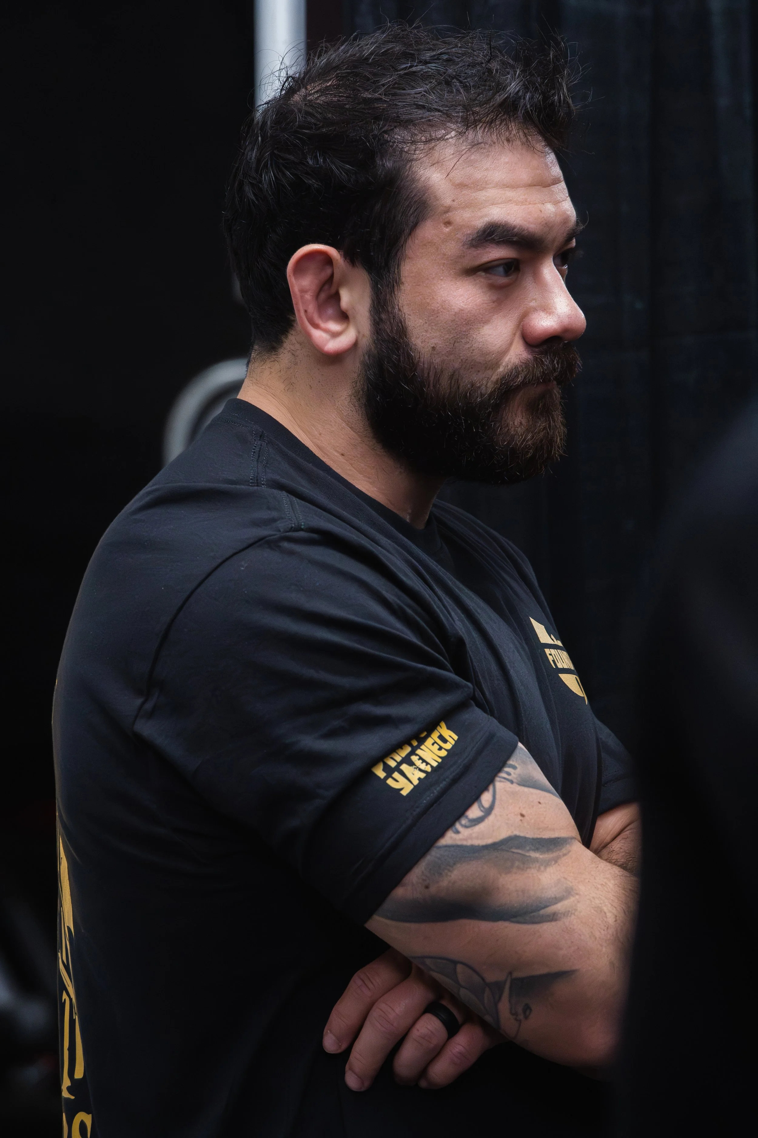 A man with a beard and dark hair standing with his arms crossed, wearing a black t-shirt with yellow text on the sleeve, in front of a dark background.