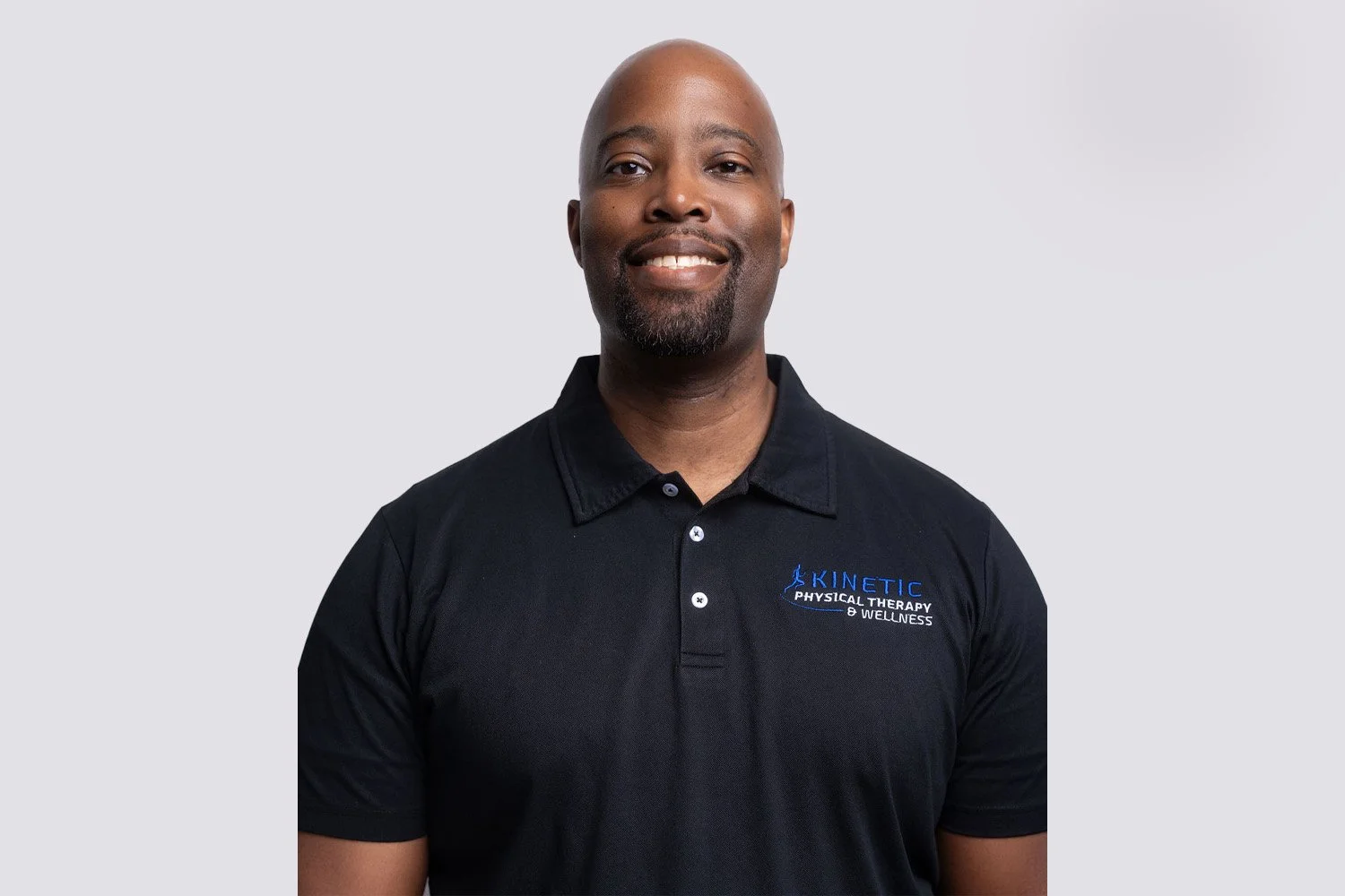 A smiling man wearing a black polo shirt with blue and white embroidered logo reading 'Kinetic Physical Therapy & Wellness' standing against a plain white background.