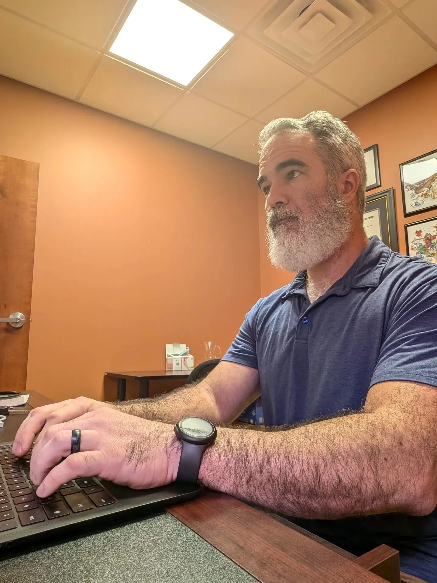 A man with gray hair and a gray beard wearing a blue polo shirt is sitting at a desk, typing on a black keyboard. The man is wearing a black watch on his left wrist. The background shows framed pictures on a peach-colored wall and a table with a tissue box.