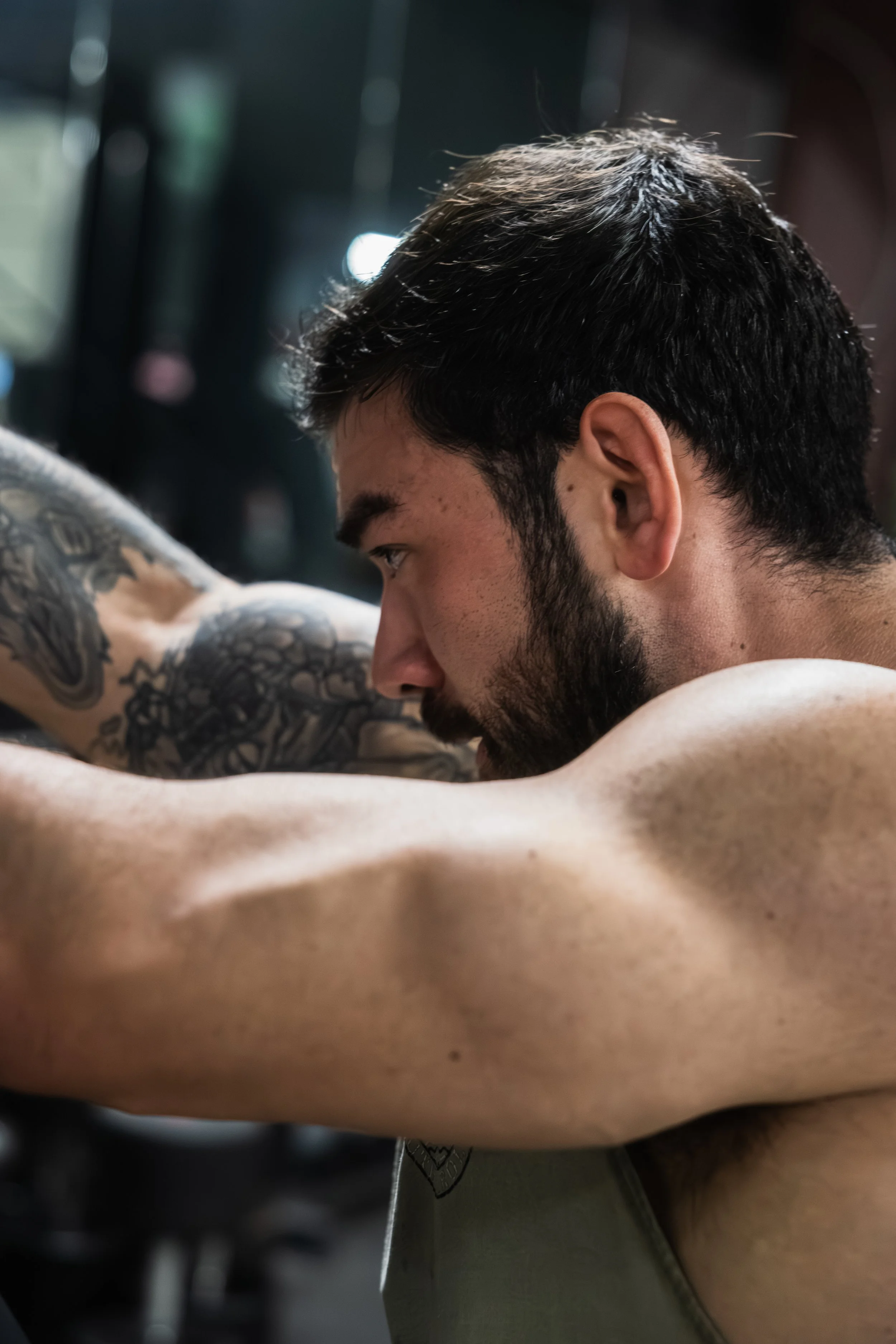 Close-up of a young man with dark hair and a beard, resting his head on his arms, with visible tattoos on his left arm, in a dimly lit indoor setting.