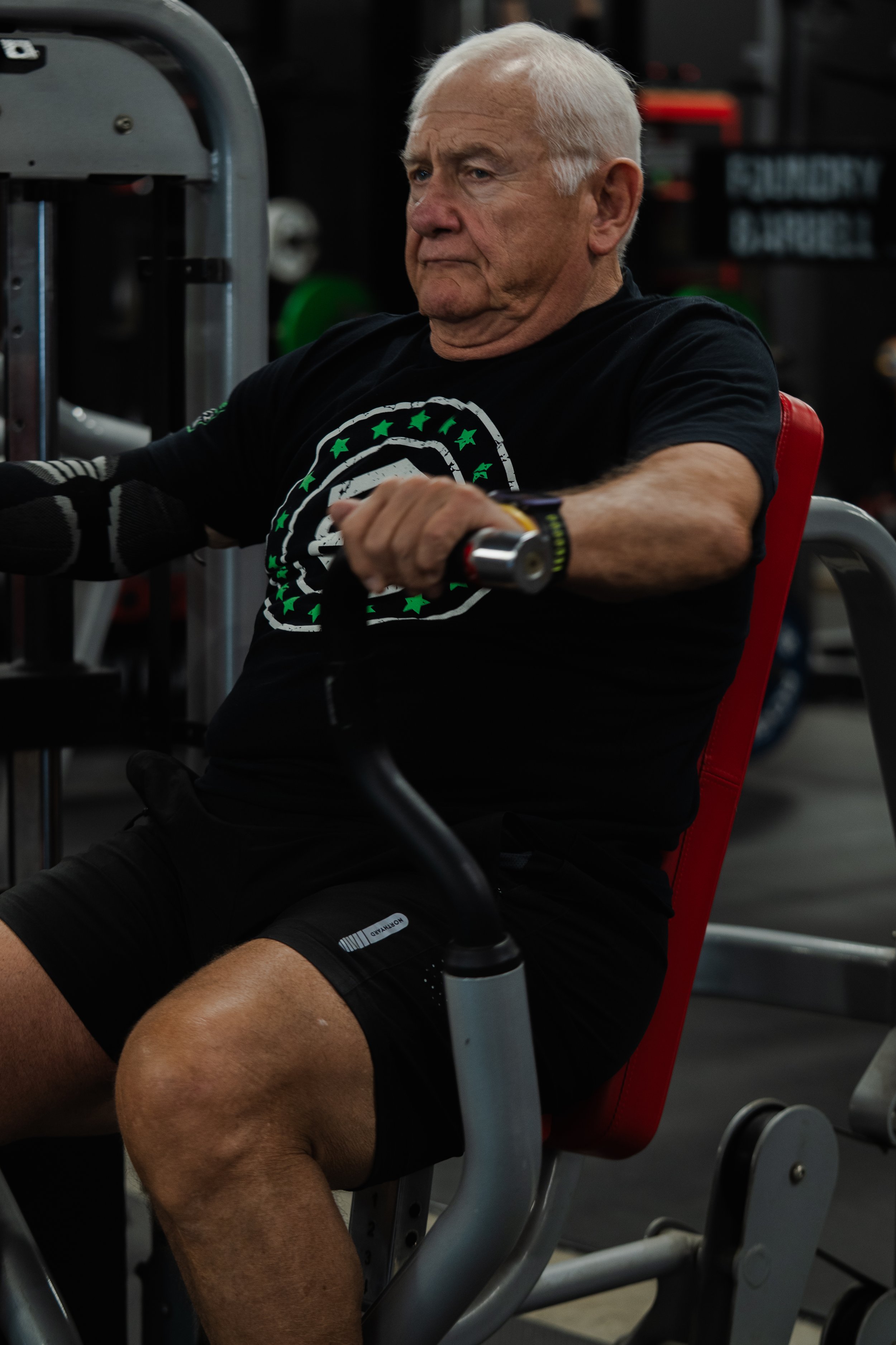 An elderly man with white hair working out on gym equipment, wearing a black T-shirt with a circular design and a wristwatch, in a gym setting with dark background and weightlifting equipment.