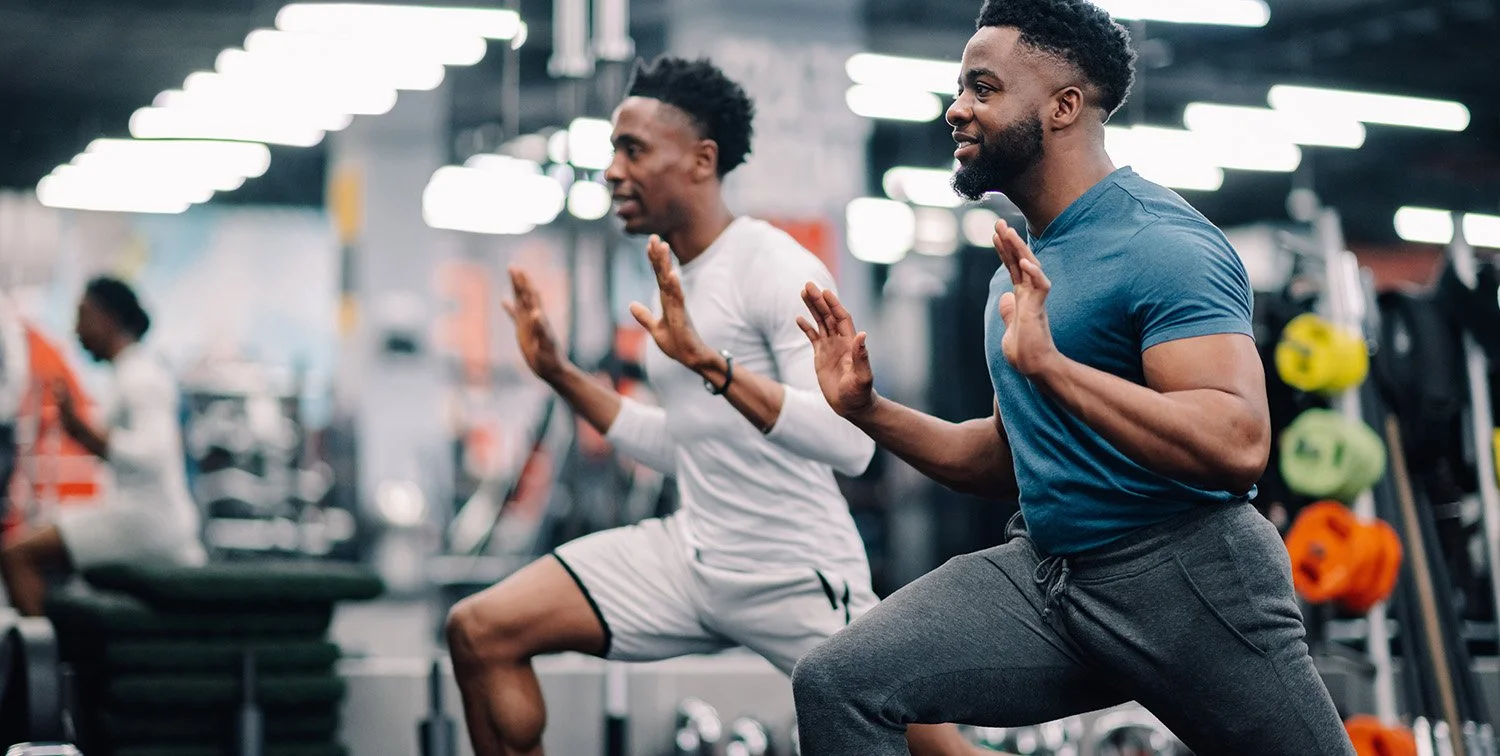 Two men doing squats in a gym, both wearing athletic clothing, with gym equipment and weights in the background.