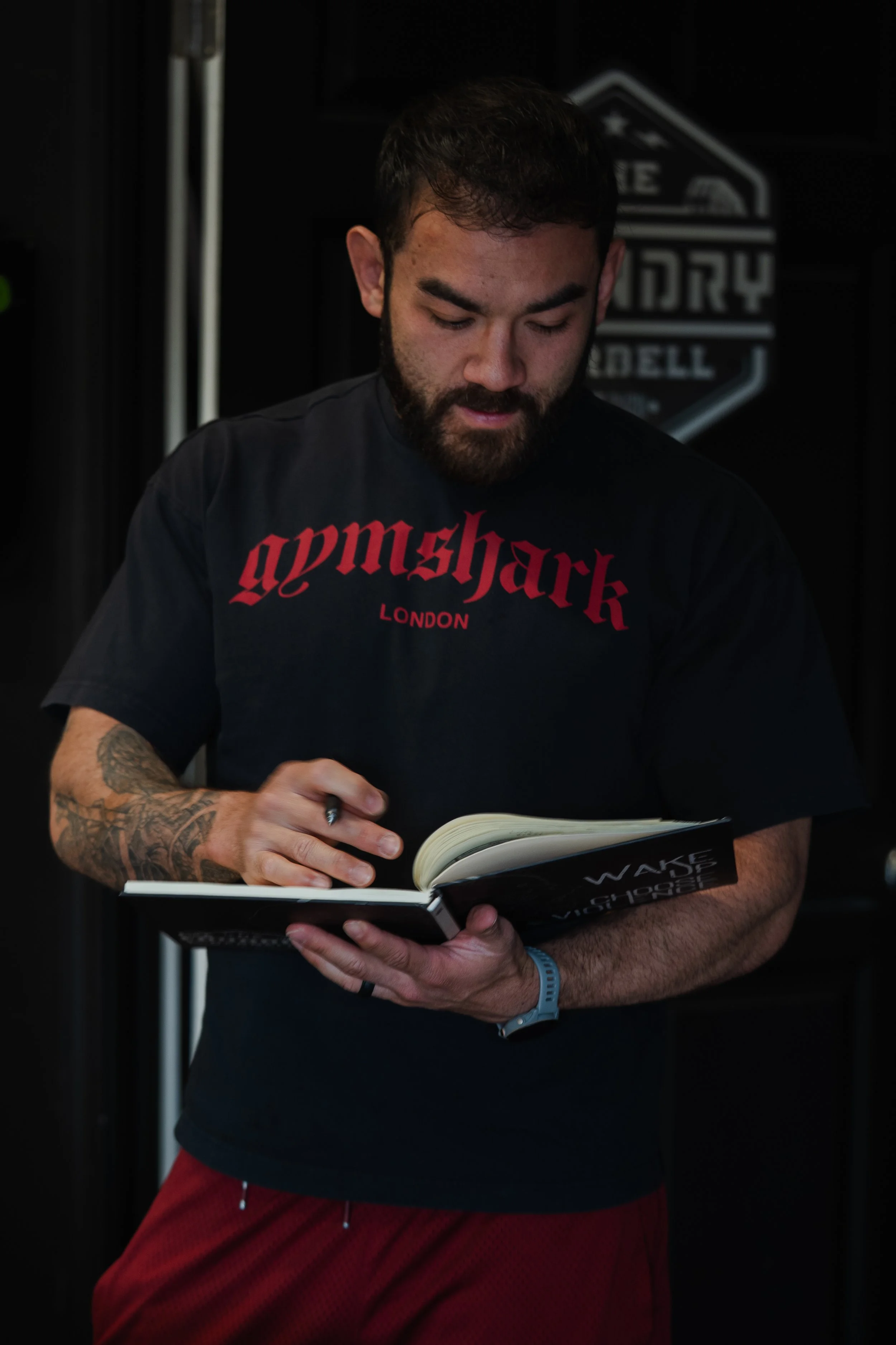 A man with a beard and tattoos on his arm, wearing a black T-shirt with red text and red shorts, is reading a book in a dark room. In the background, there is a sign with the words "WAKE UP, VISION, CHOPPER".