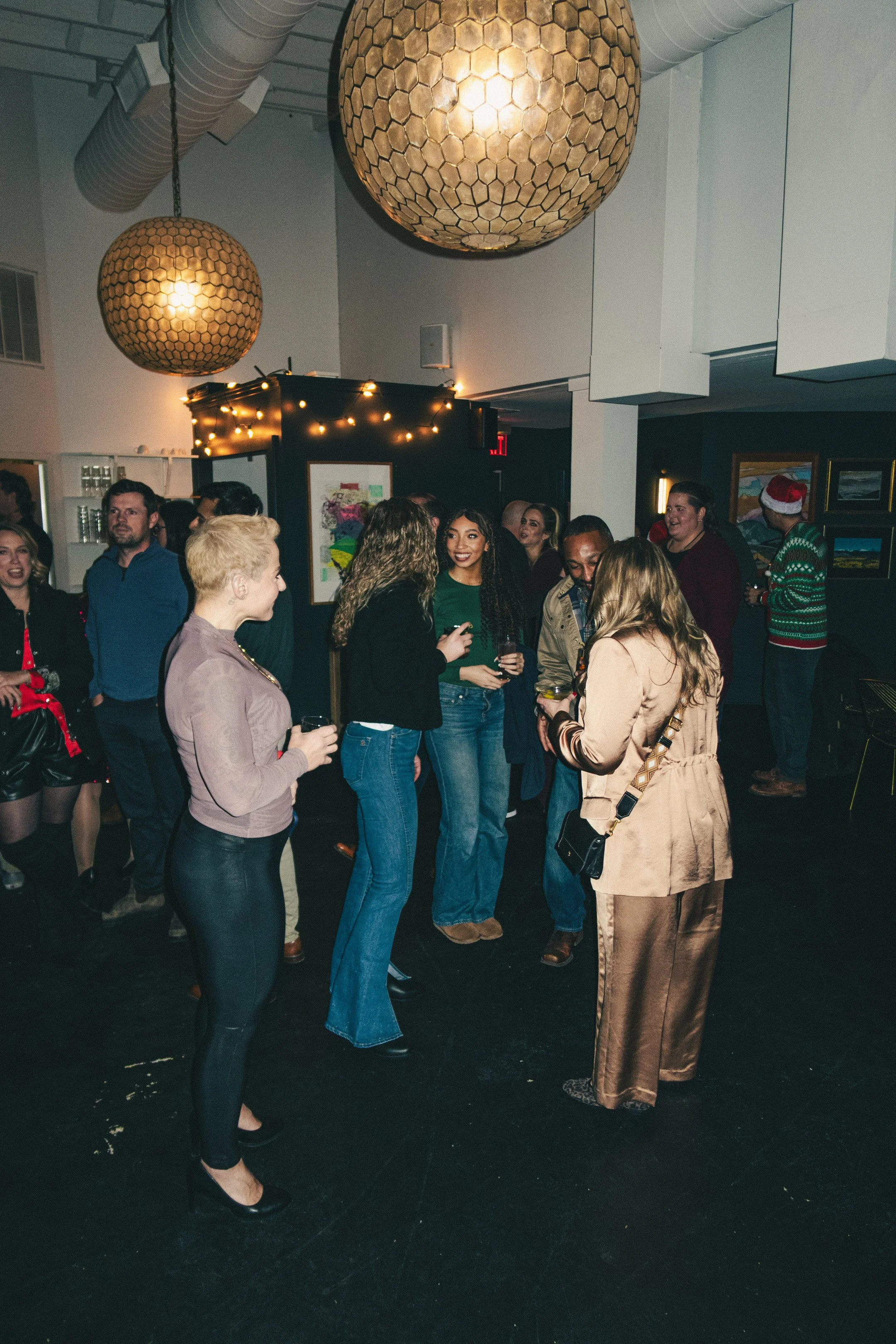 A group of people socializing at an indoor party with modern decor, large hanging lights, and framed artwork on walls. Some are holding drinks and wearing casual to semi-formal clothing.