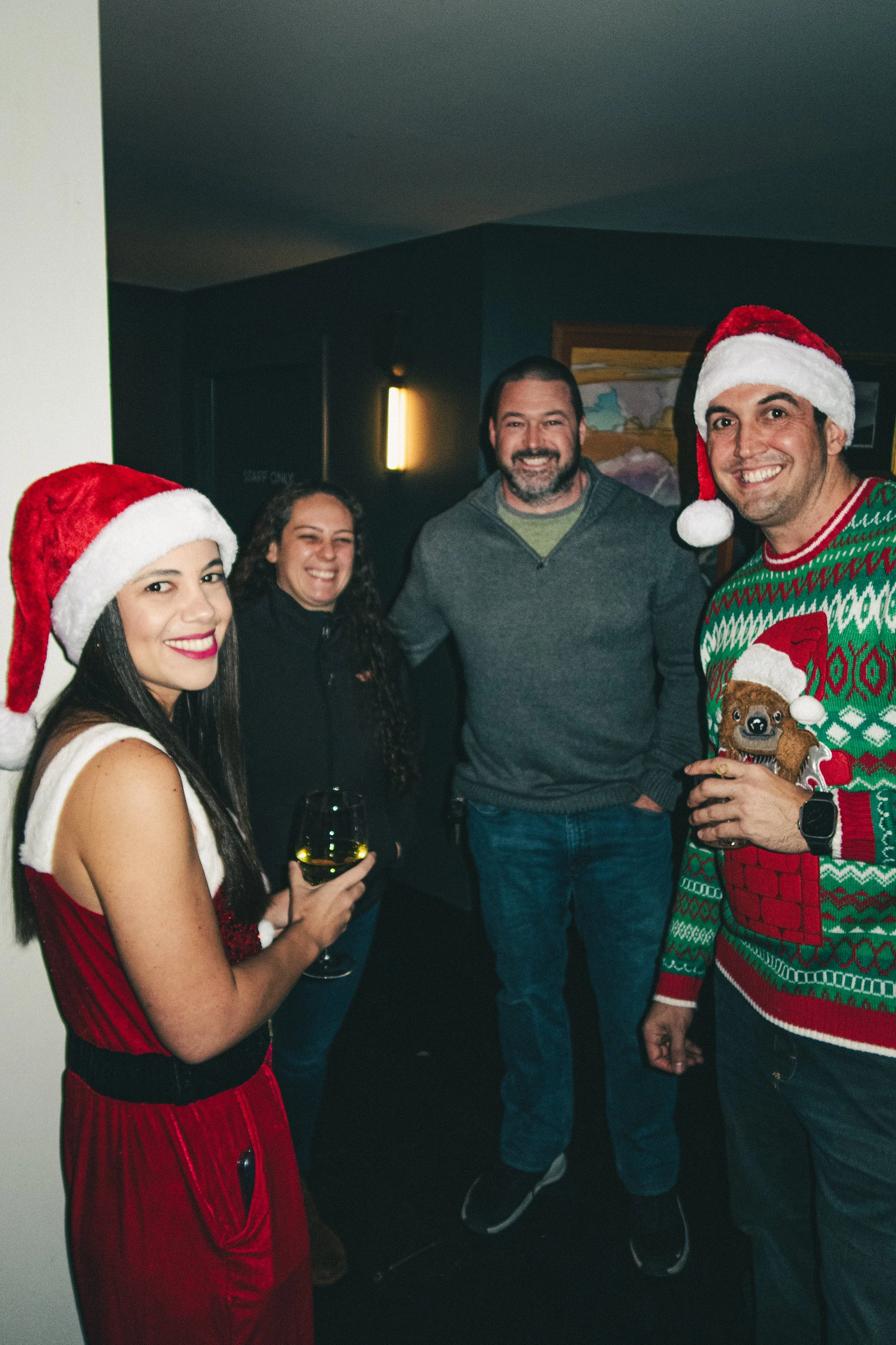 Group of four adults celebrating Christmas indoors, with two women wearing Santa hats and holding drinks, and two men, one wearing a Christmas sweater and Santa hat, smiling and posing for the photo.