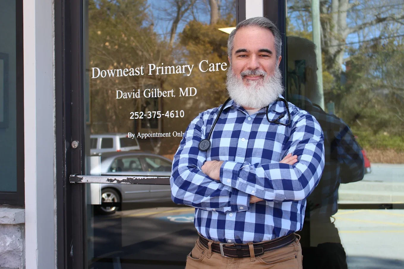 A man with a gray beard and gray hair standing outside a medical office, wearing a blue plaid shirt and a stethoscope around his neck, with arms crossed, smiling.