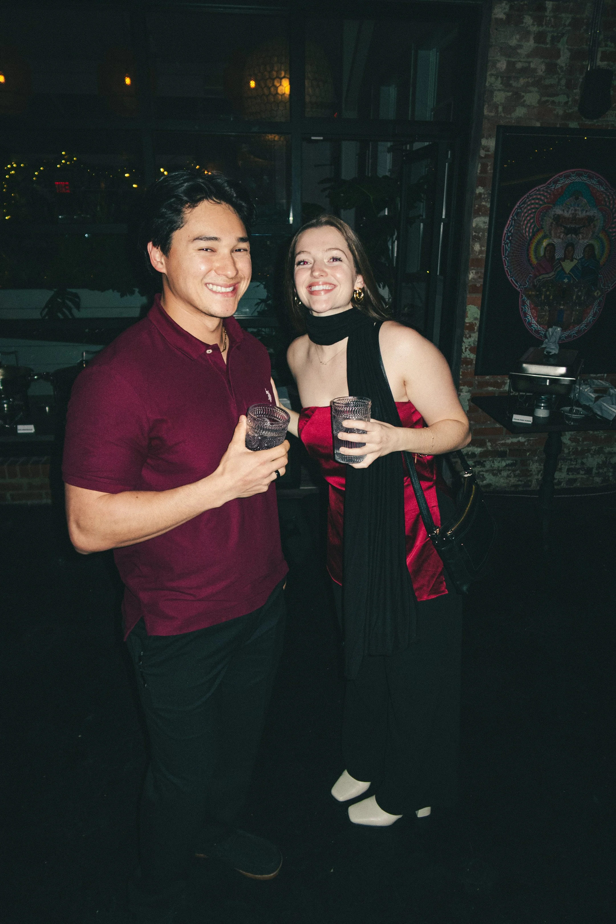 A young man and woman smiling and holding drinks at a party or social gathering indoors with a brick wall, window, and artwork in the background.