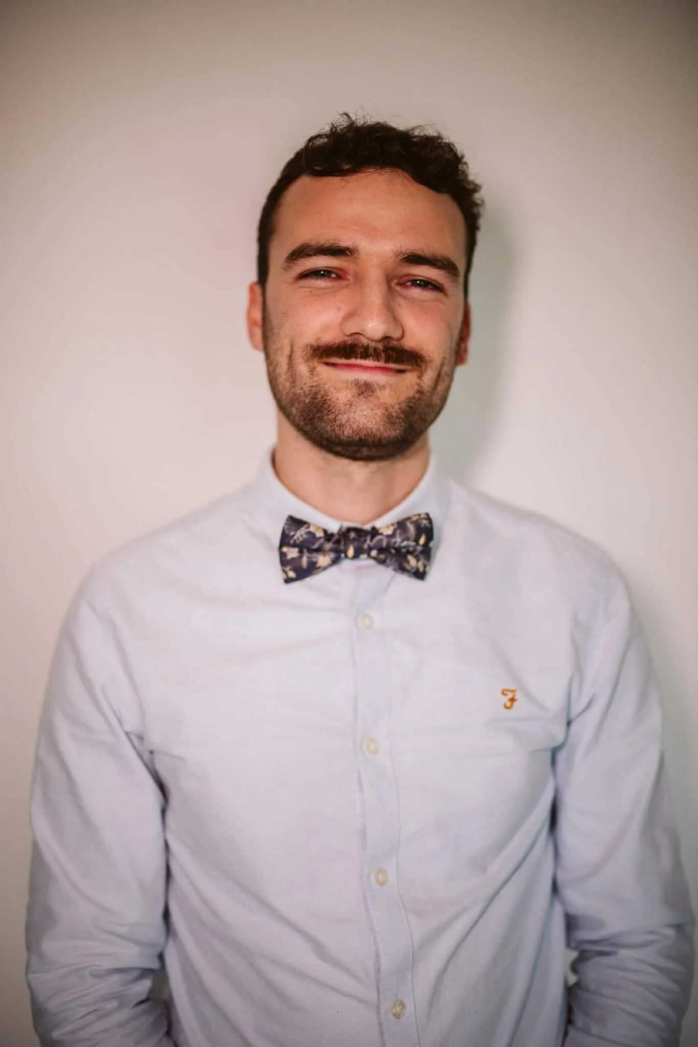 Jeune homme souriant portant une chemise blanche et un nœud papillon assis contre un mur blanc.