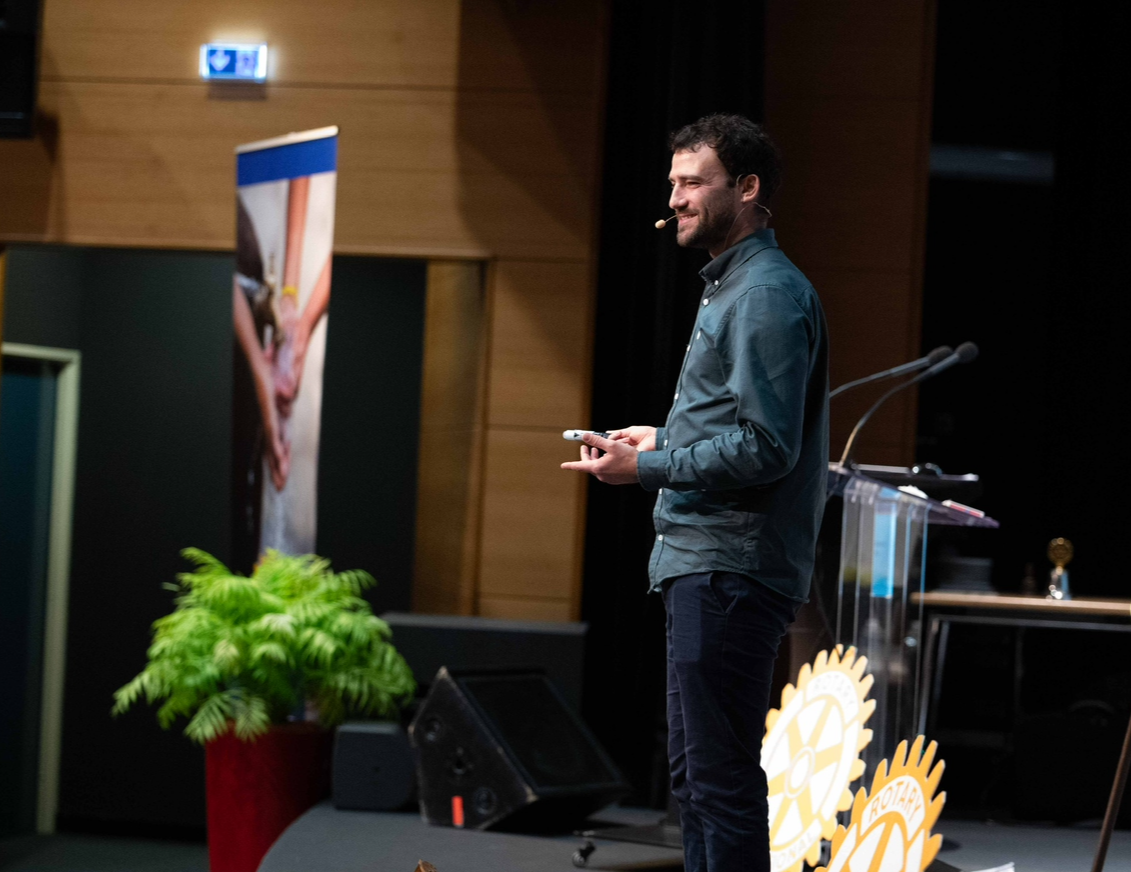 Un homme parle lors d'une conférence, debout sur scène avec micro-casque, fond en bois et pupitre avec symbole de Rotary