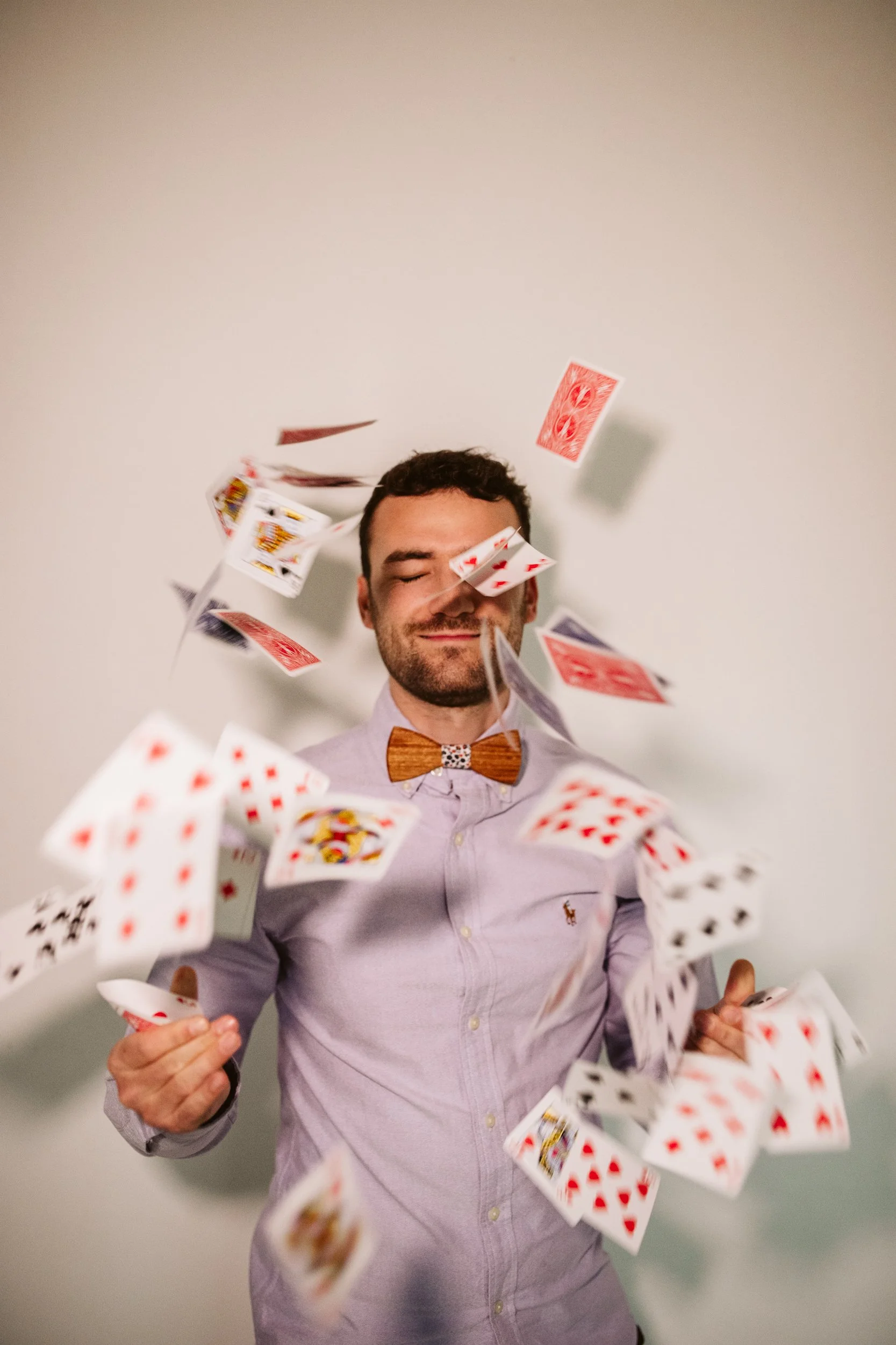 Un homme souriant avec un nœud papillon en bois, portant une chemise lilas, entouré de cartes à jouer en suspension dans l'air
