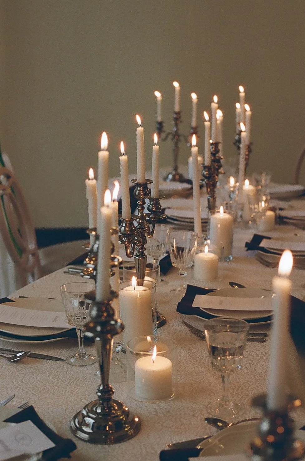 A long dining table set for a reception, illuminated by numerous glowing white taper candles in silver candelabras, creating a warm and intimate atmosphere.