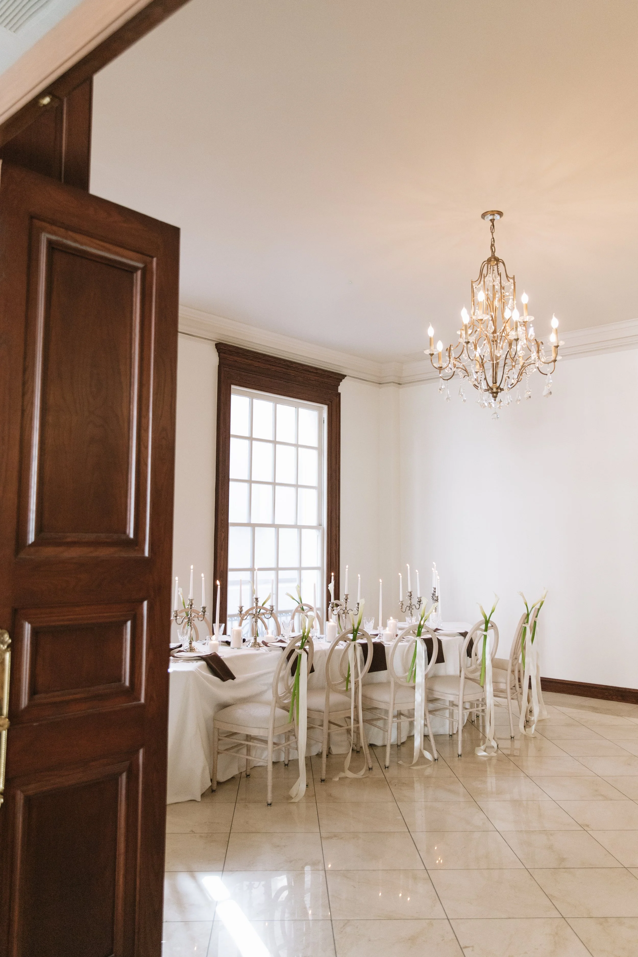 A wide shot framed by an open dark wood double door, showing a long reception table set with white linens, lit crystal chandeliers, and matching chairs decorated with white calla lilies.