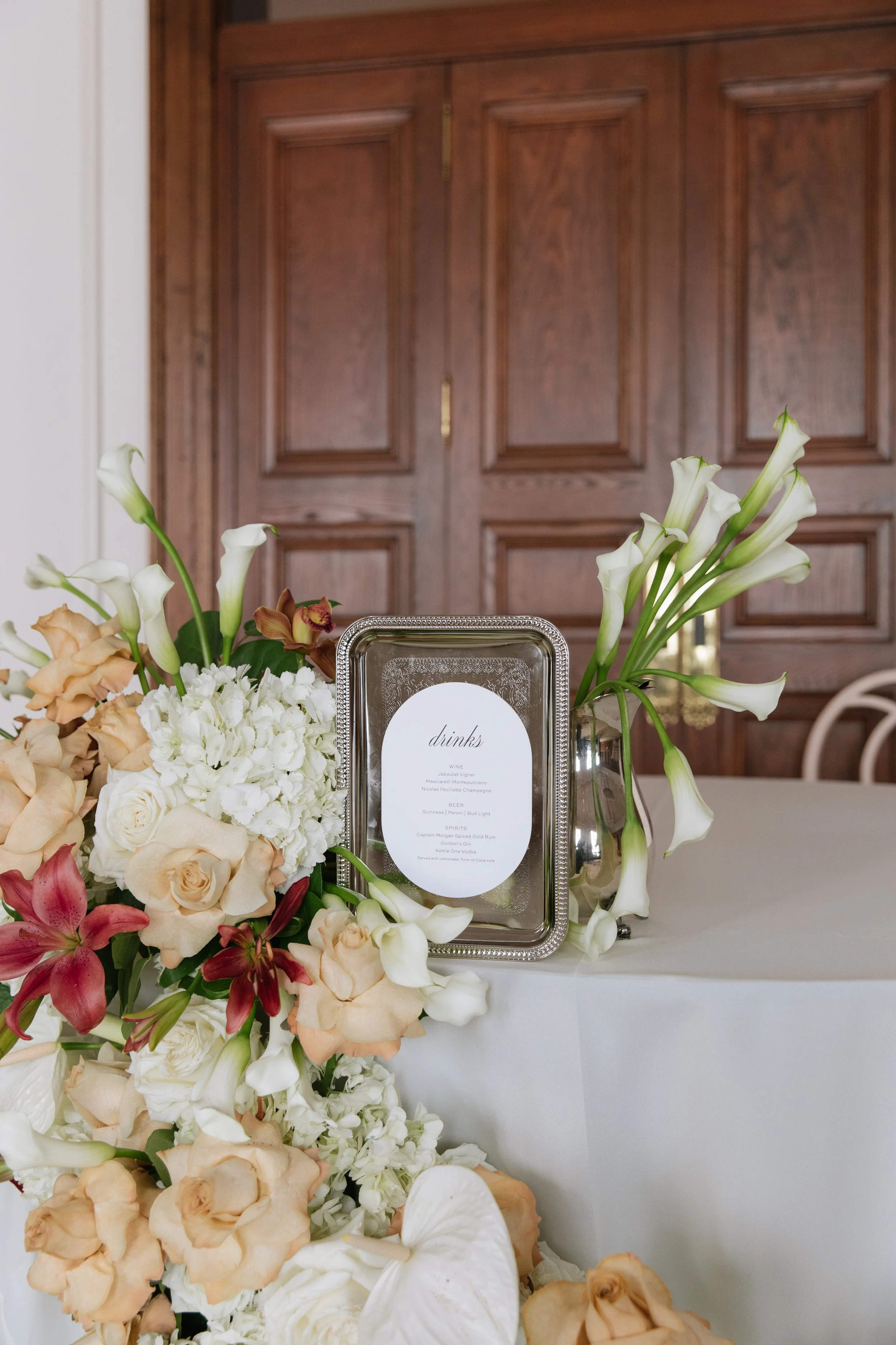 An oval drink menu resting on an intricate silver tray, framed by a vibrant floral arrangement of white hydrangeas, roses, and calla lilies against a dark wood paneled wall.