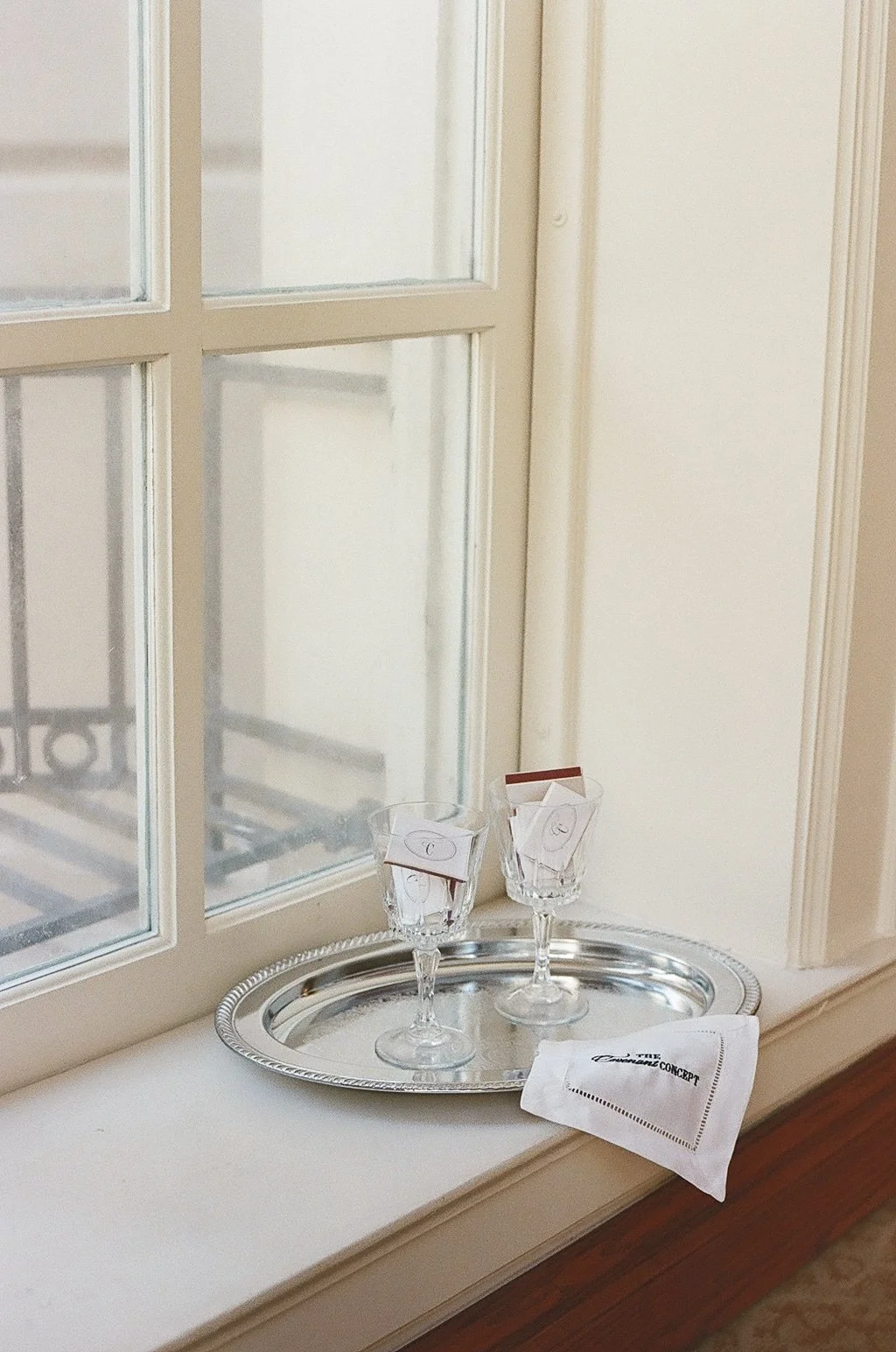 Two wine glasses and an embroidered white napkin placed on a silver tray by a window sill.
