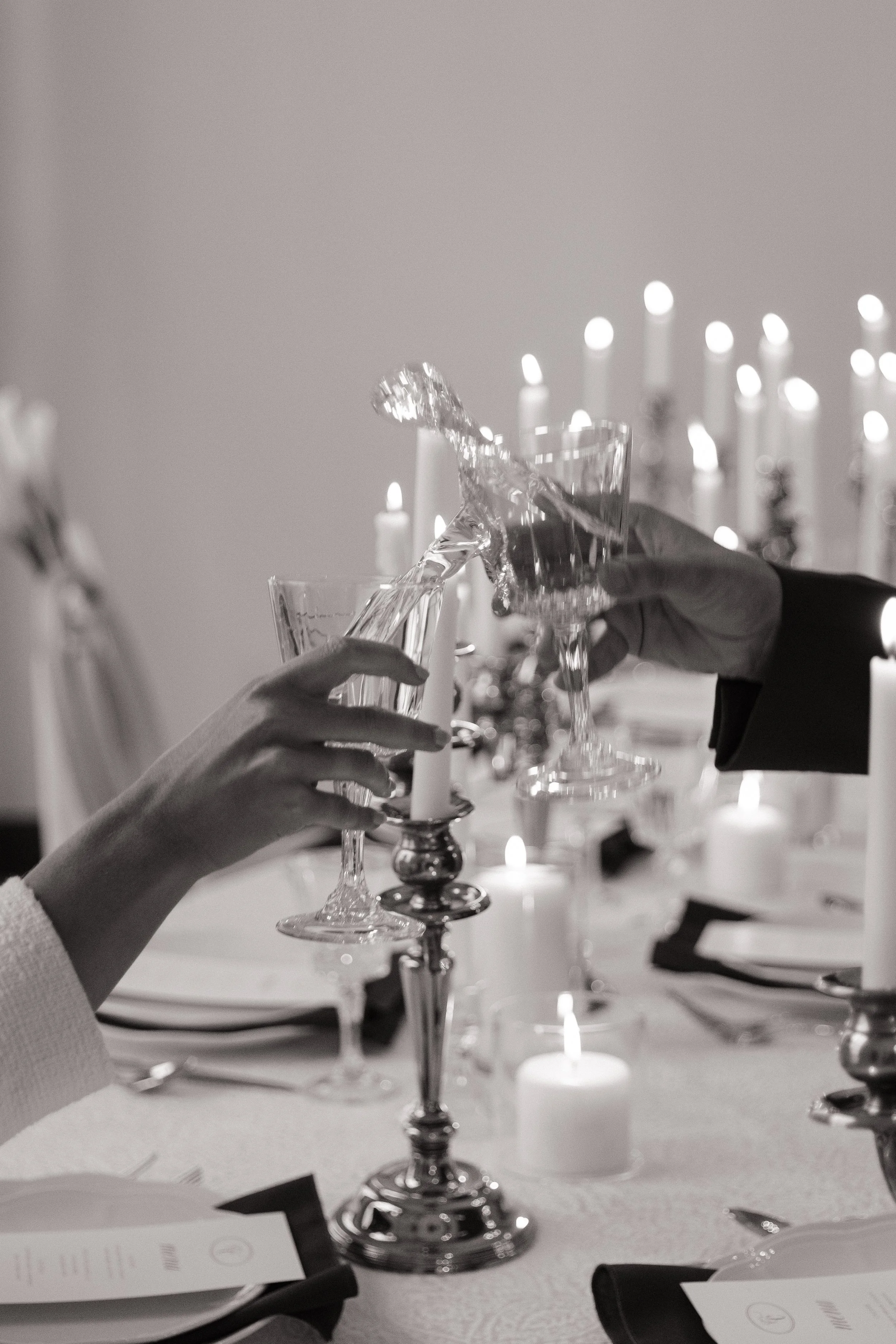 A black and white close-up photograph of a couple clinking crystal wine glasses over a candlelit reception table.
