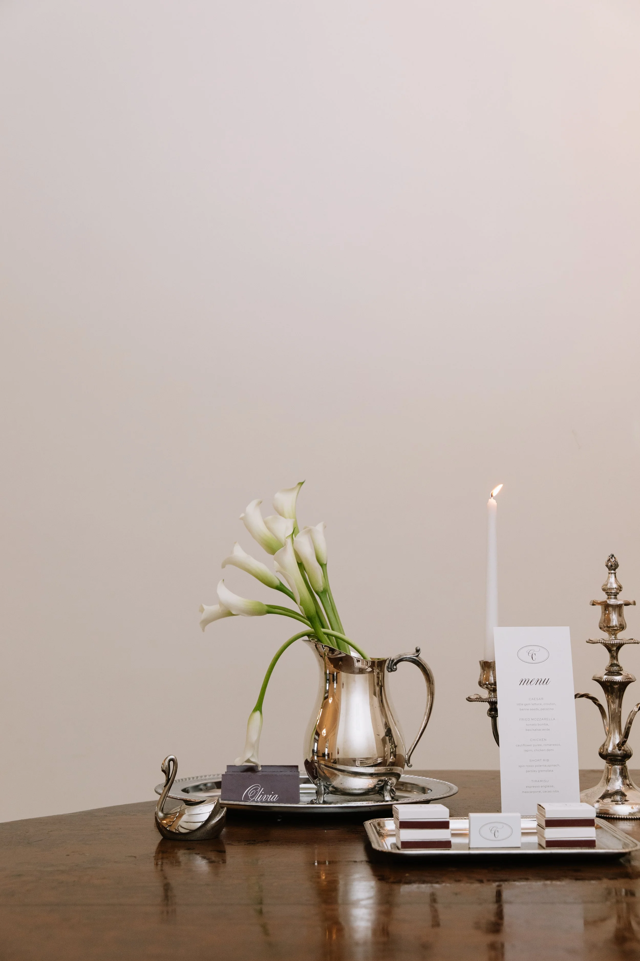 A silver pitcher holding white calla lilies sits on a dark wood table next to a menu card and lit taper candles.