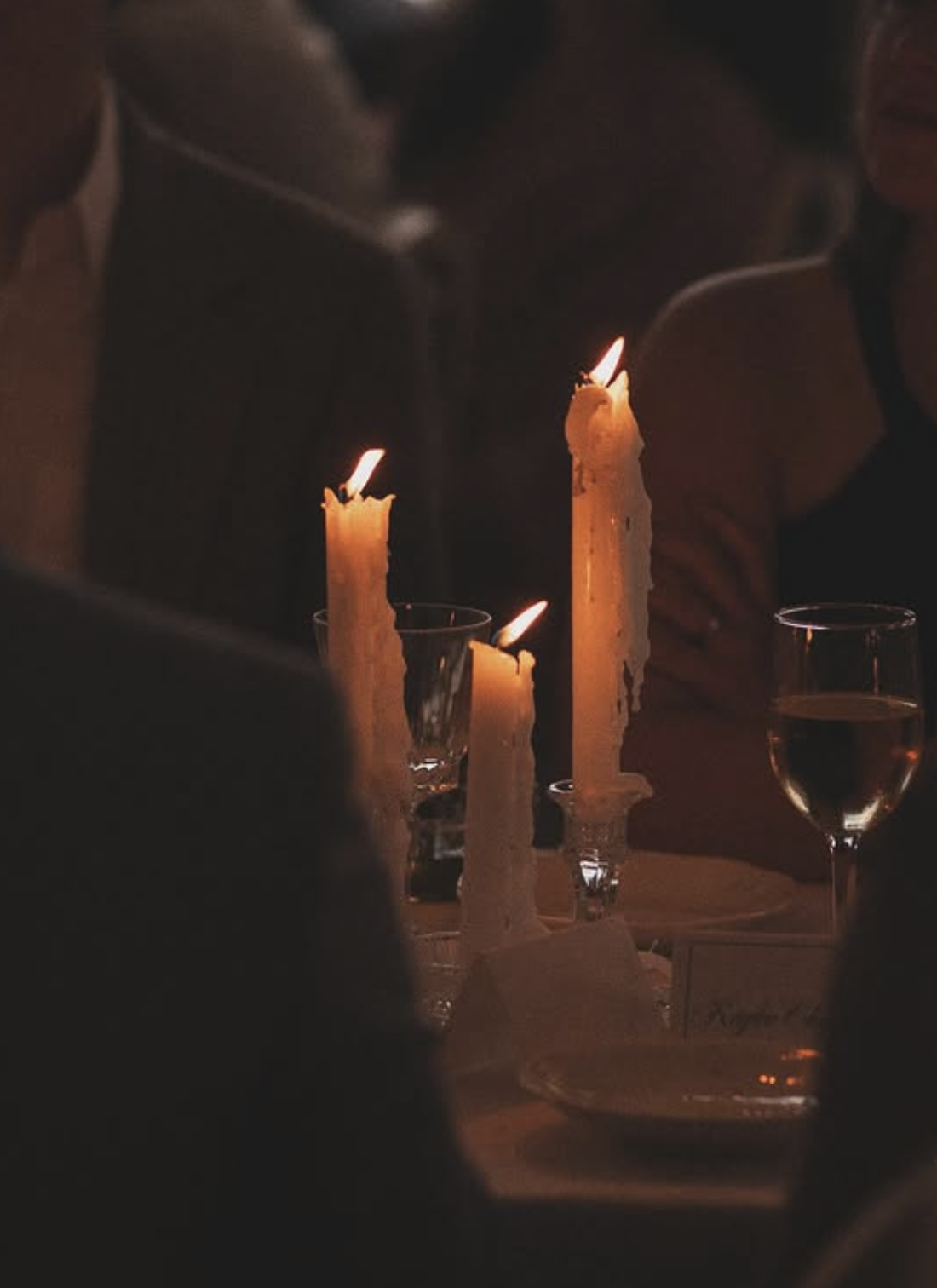 A close-up, warm-toned view of tall, dripping white taper candles glowing on a reception dinner table with wine glasses.