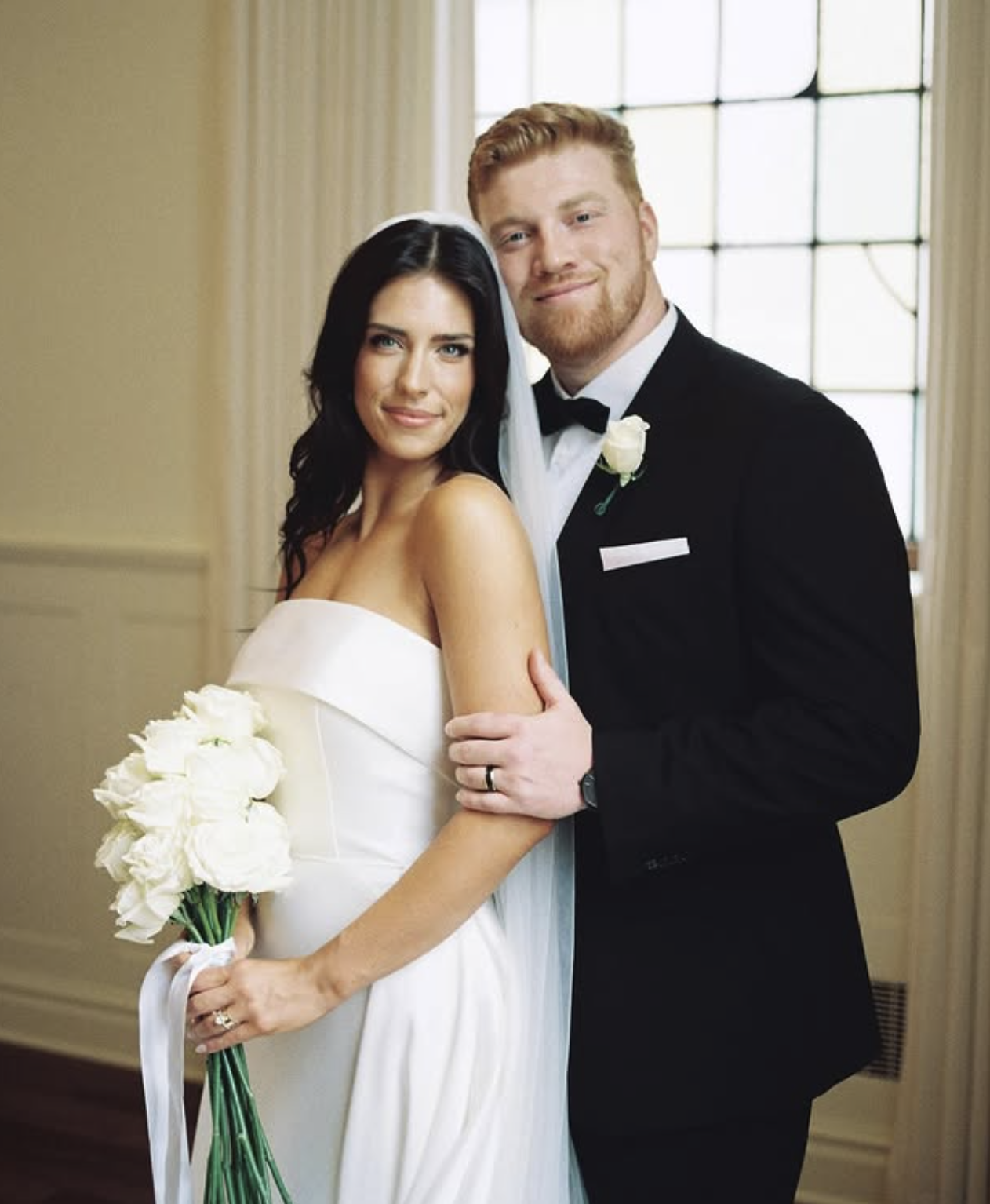 A bride in a strapless white wedding dress and veil stands with a groom wearing a classic black tuxedo, holding a tight bouquet of white roses.