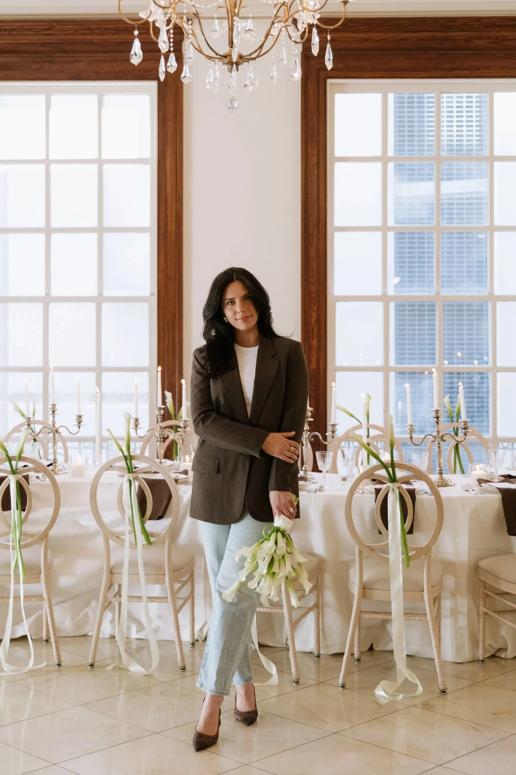 A woman in a brown blazer, white t-shirt, and blue jeans stands in front of a long, white-clothed reception table lined with lit candelabras and white calla lilies, holding a matching bouquet.