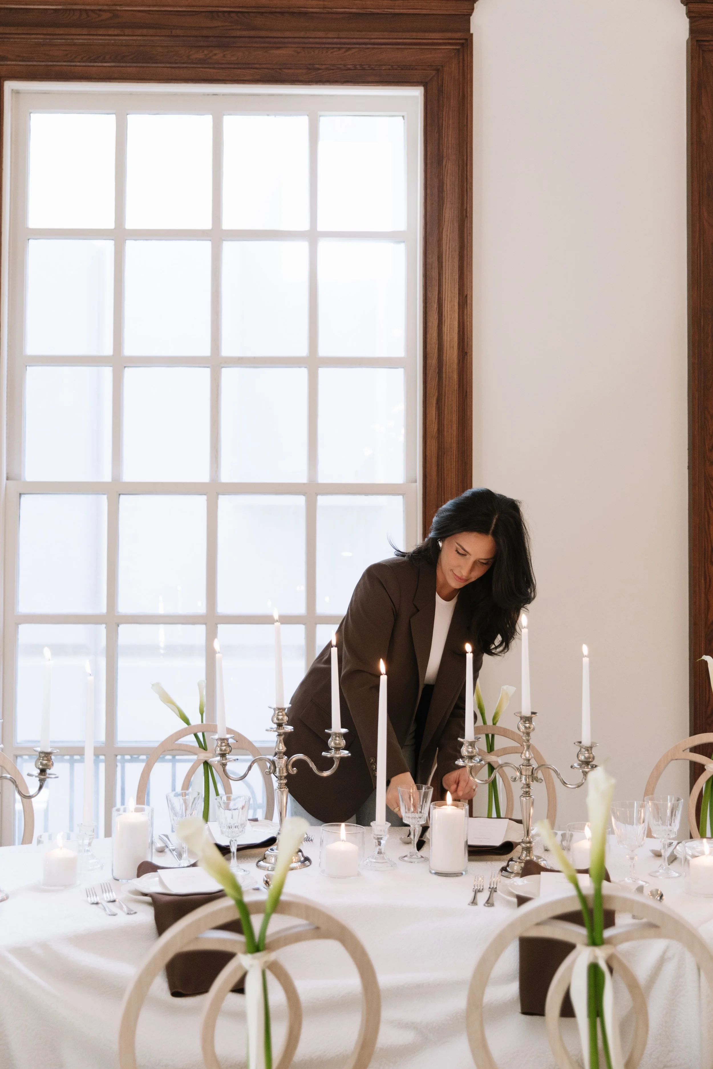 A woman in a brown blazer adjusts lit white taper candles on a silver candelabra set along a reception table by a large window.