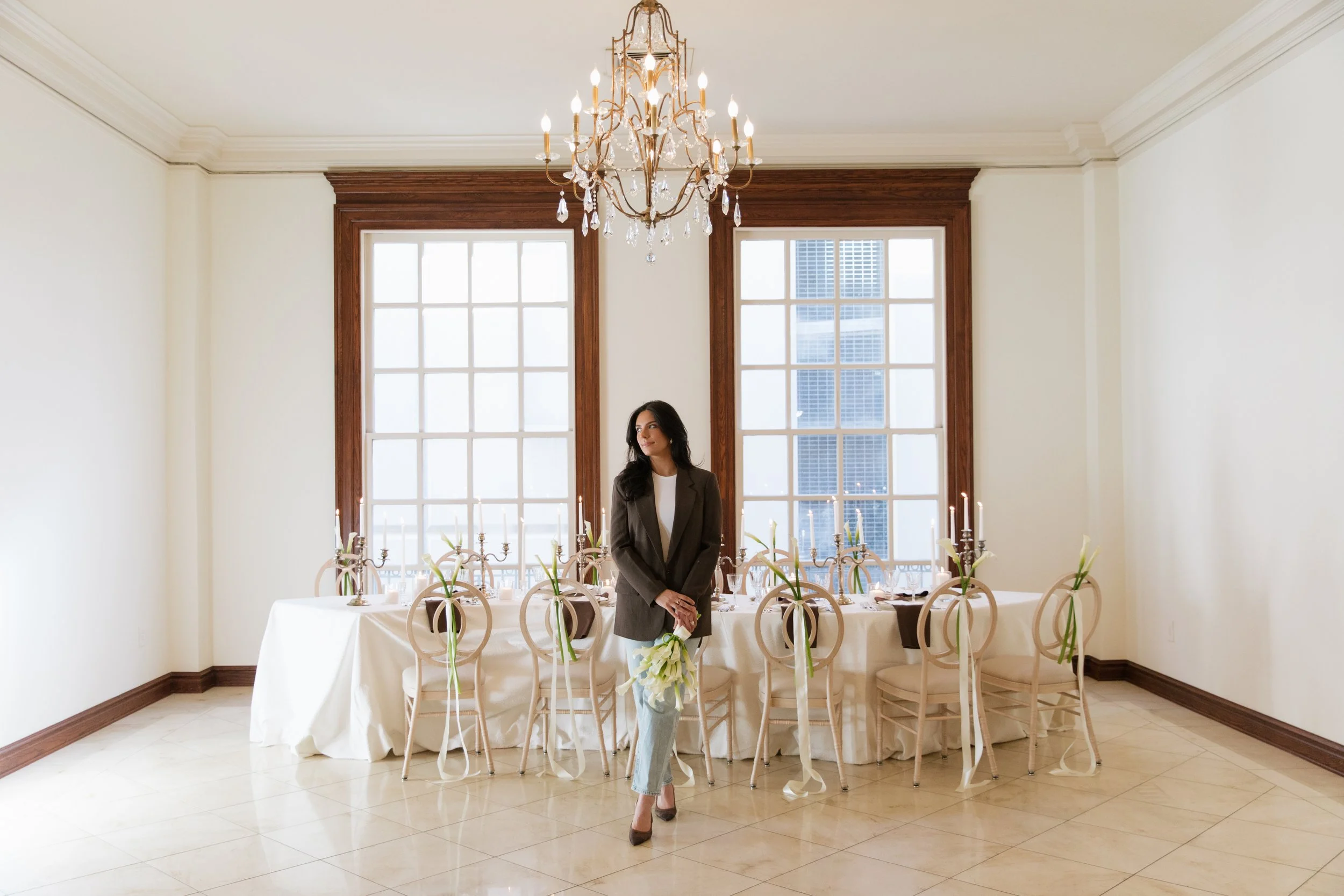 A woman in a brown blazer and jeans stands holding a bouquet of white calla lilies, centered in front of a long, beautifully set wedding reception table between two large windows.
