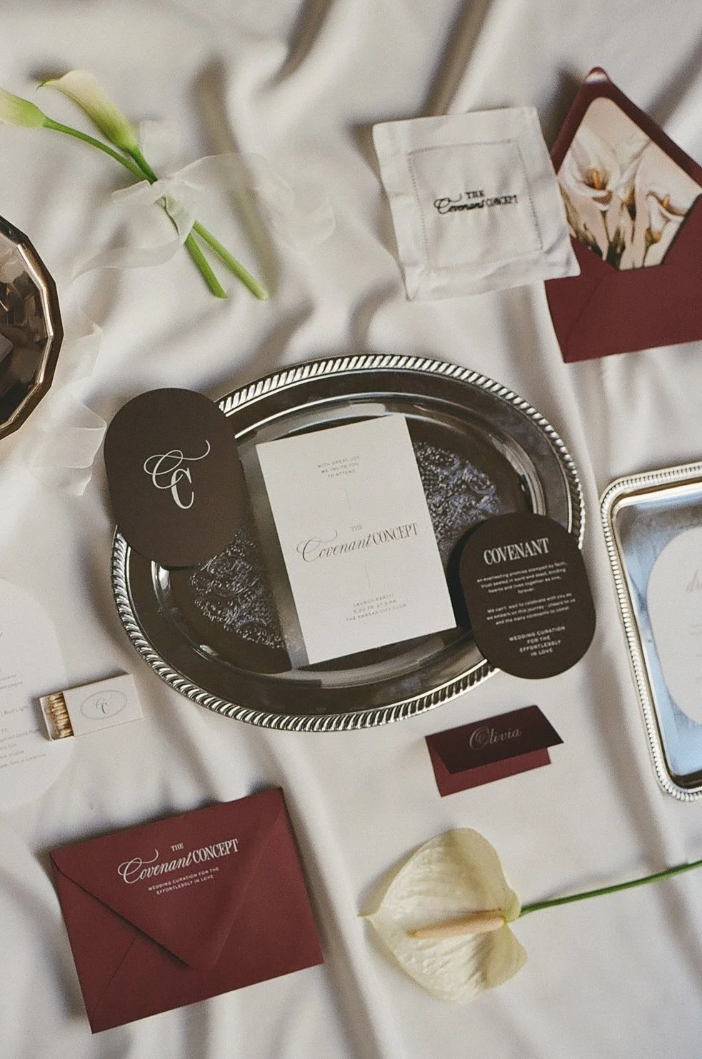 An overhead flat lay of a wedding stationery suite featuring an invitation on a silver tray, surrounded by dark burgundy envelopes and delicate white floral accents.