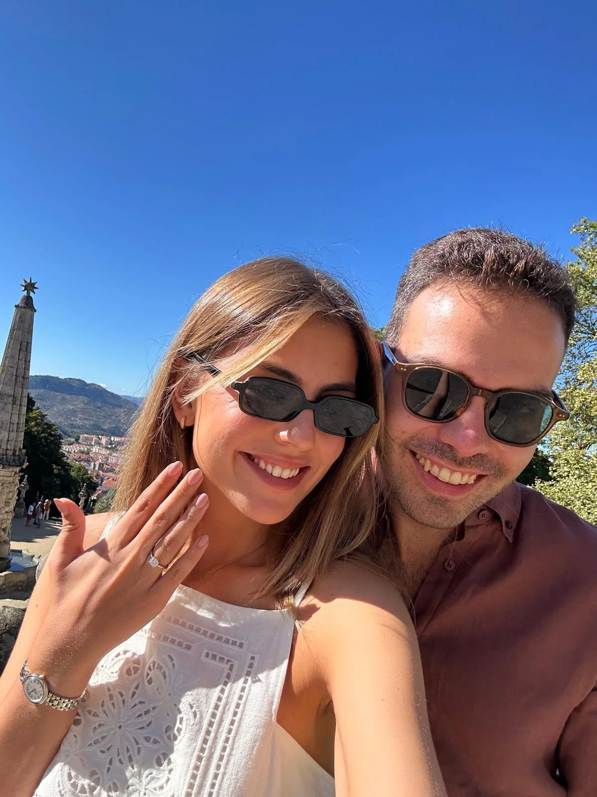 Casal sorridente usando óculos escuros tira selfie ao ar livre com céu azul claro.