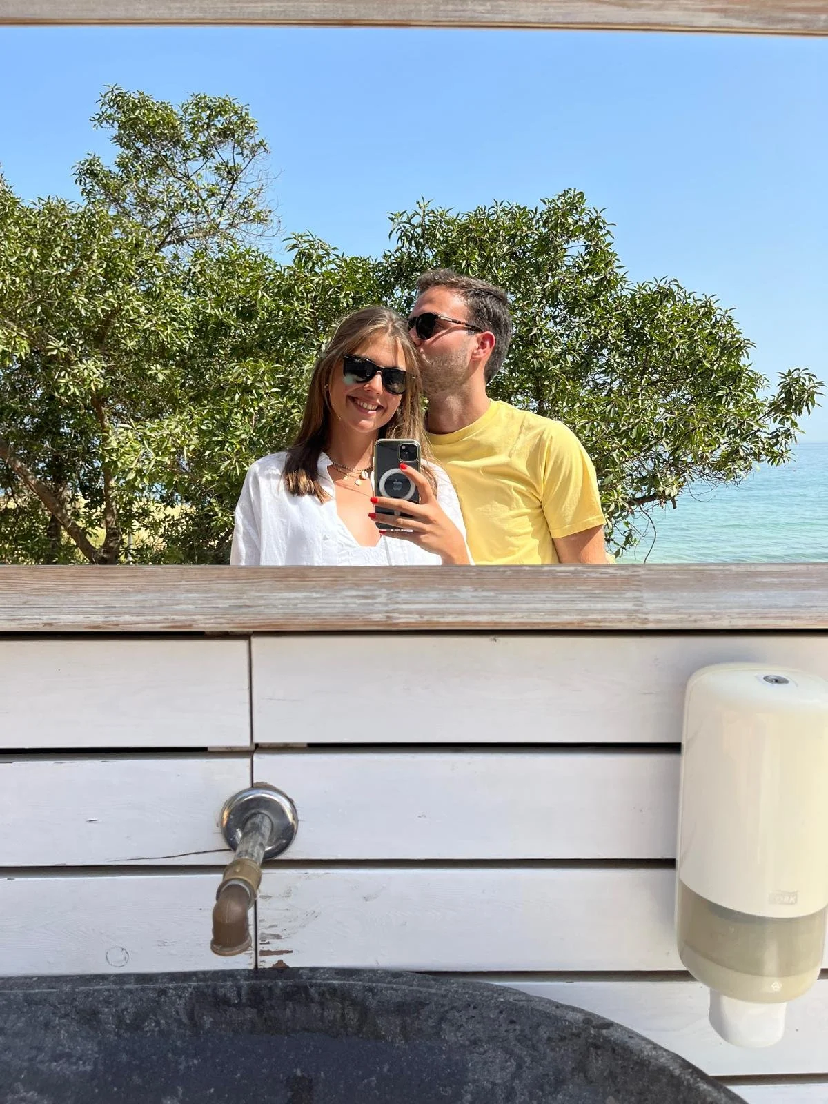 Um casal tira uma selfie em um espelho ao ar livre, com árvores verdes e céu azul ao fundo.