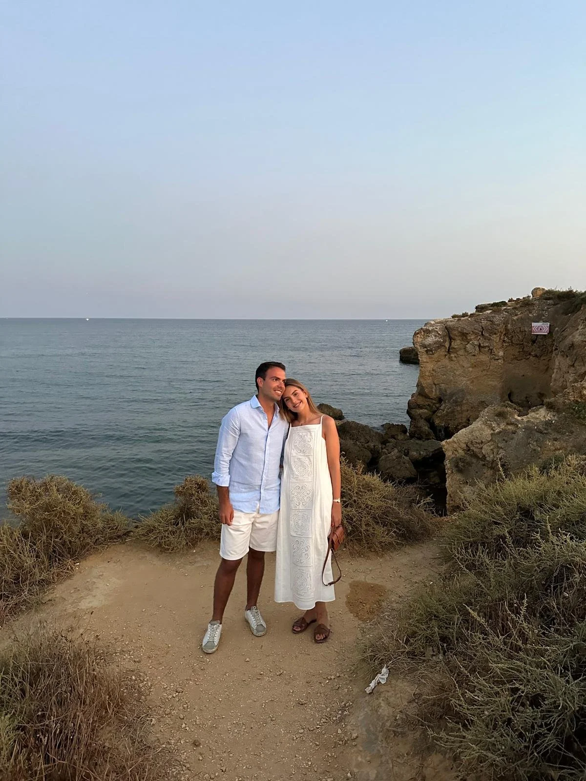 Um casal sorridente na praia, com o mar ao fundo, ao pôr do sol, vestidos com roupas claras, caminhando próximo às rochas.