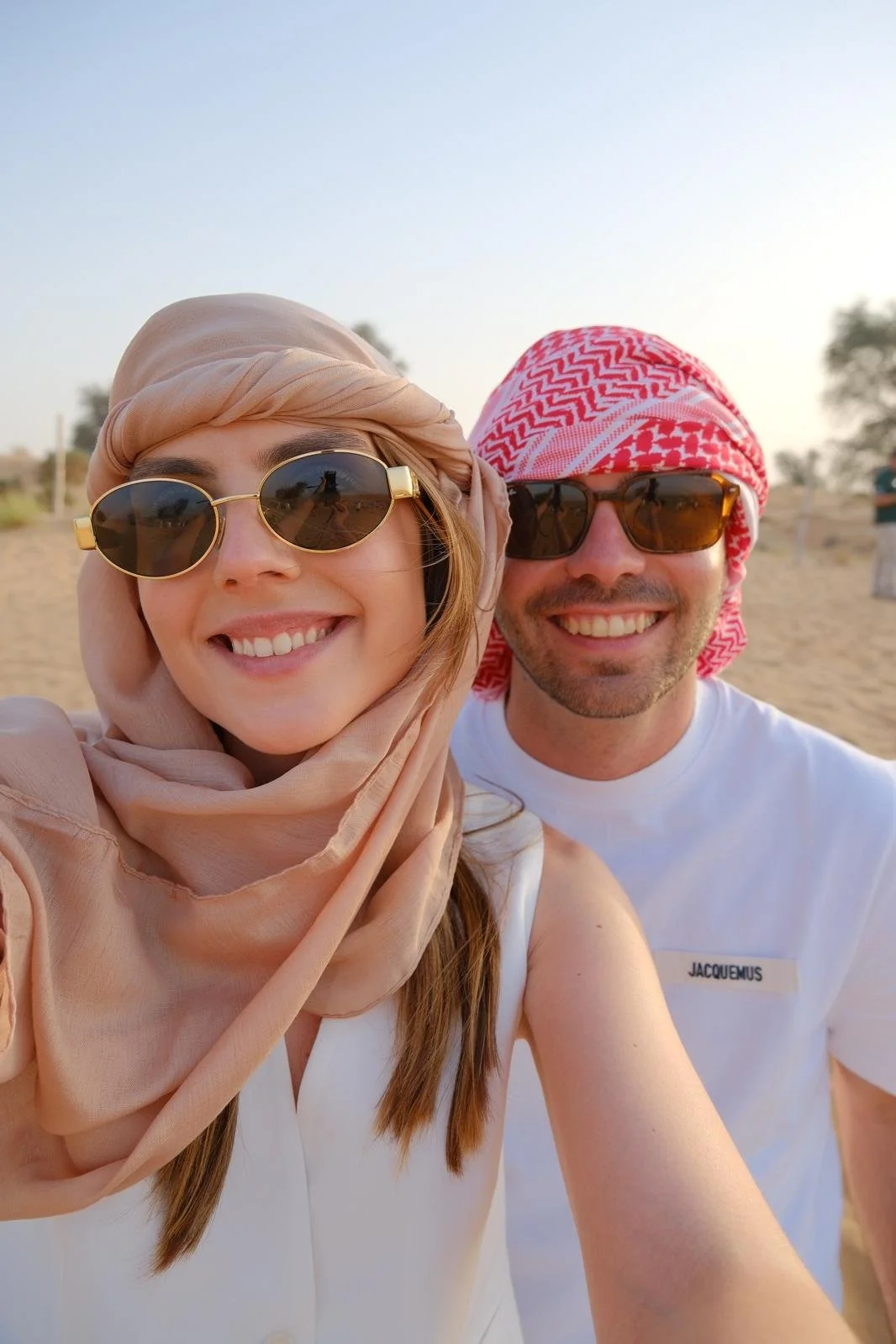 Casal sorridente usando óculos de sol e chapéus, posando em uma praia ao pôr do sol.