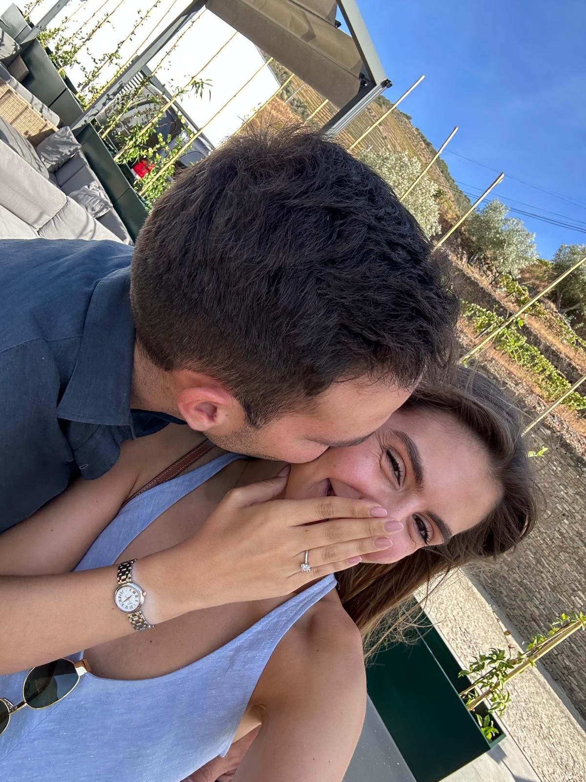 Um homem beijando uma mulher na bochecha ao ar livre, enquanto ela cobre a boca com a mão, sorrindo. Ela usa um vestido cinza claro, um relógio no pulso esquerdo e um anel de noivado na mão esquerda.