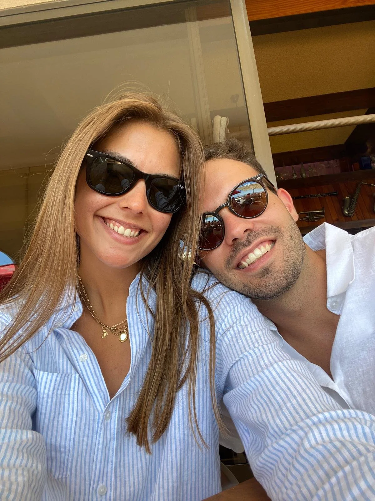 Um casal sorridente usando óculos de sol, posando para uma selfie em um ambiente ao ar livre.