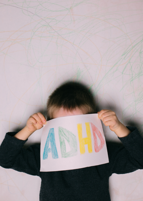 Child holding a drawing with the letters 'ADHD' written in colorful markers, covering their face, against a wall with scribbles.