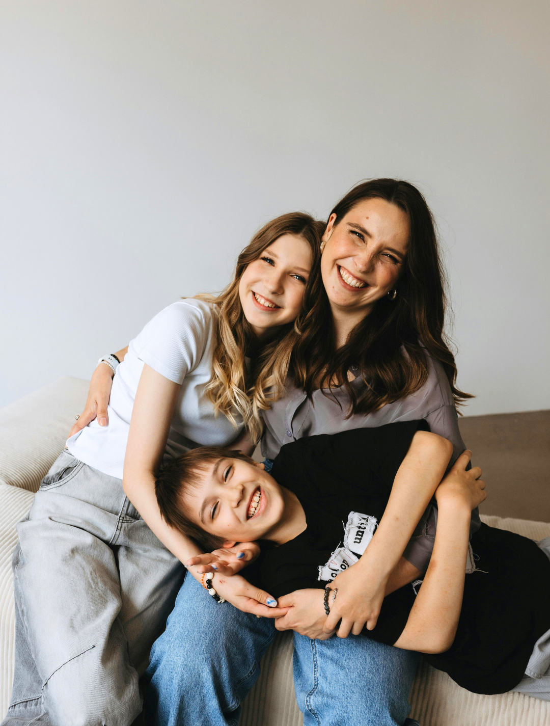 Three people, a woman and two children, smiling and hugging on a couch in a well-lit room.