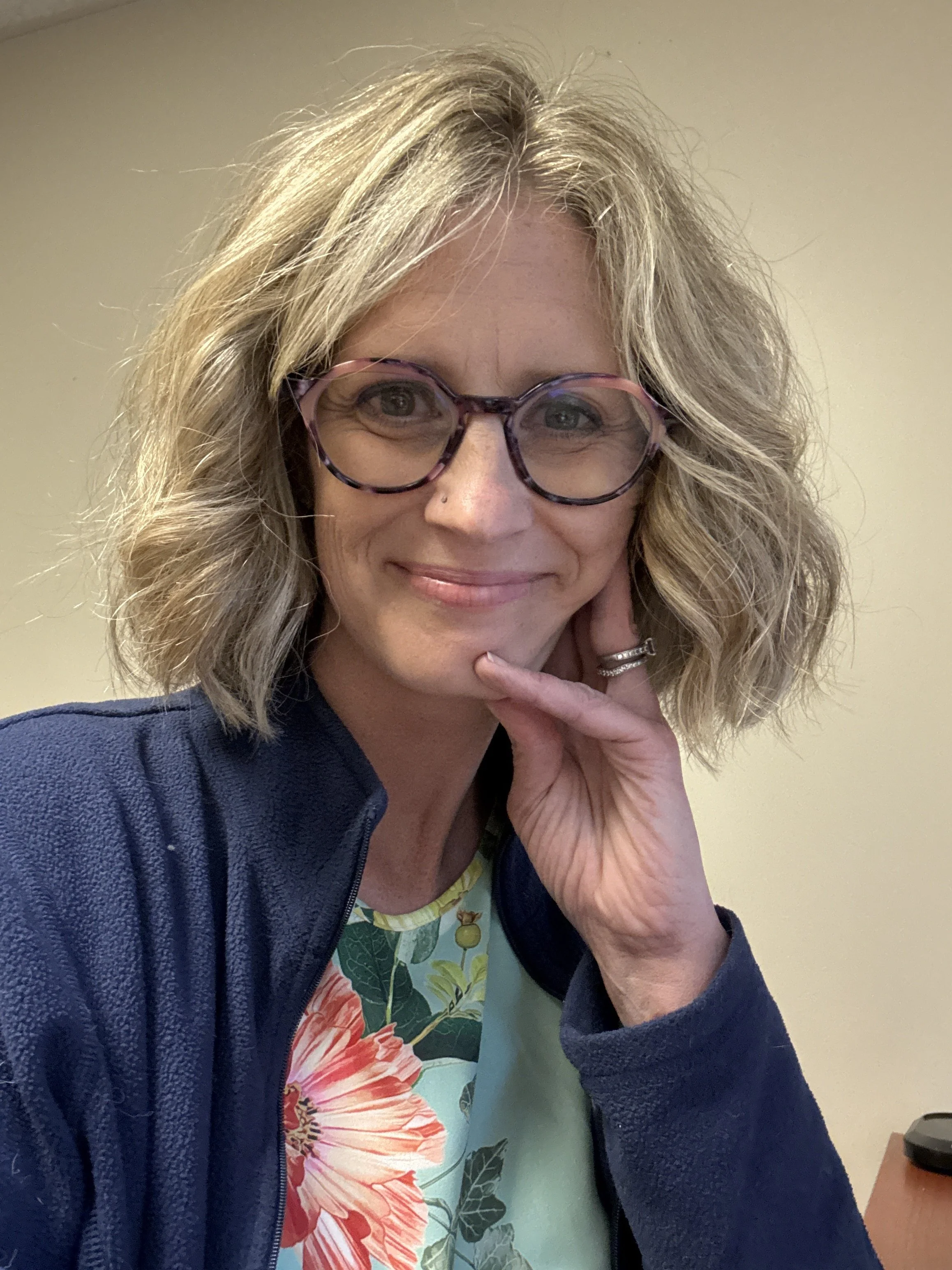 A woman with shoulder-length blonde hair, wearing glasses, a dark blue jacket, and a floral shirt, smiling and holding her hand to her chin.
