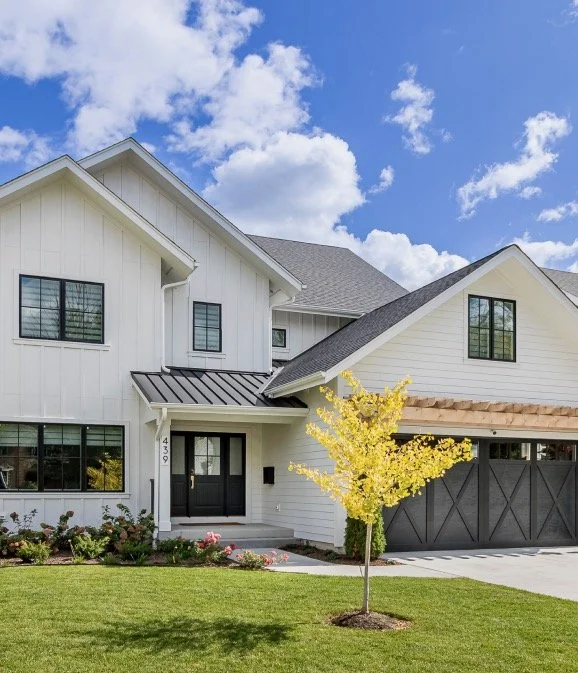 Front view of a modern two-story white house with black window frames, a black front door, and a two-car garage with a dark gray door. There is a small yellow-leafed tree in the front yard, with a well-manicured green lawn and some flower bushes. The sky is partly cloudy with white clouds.