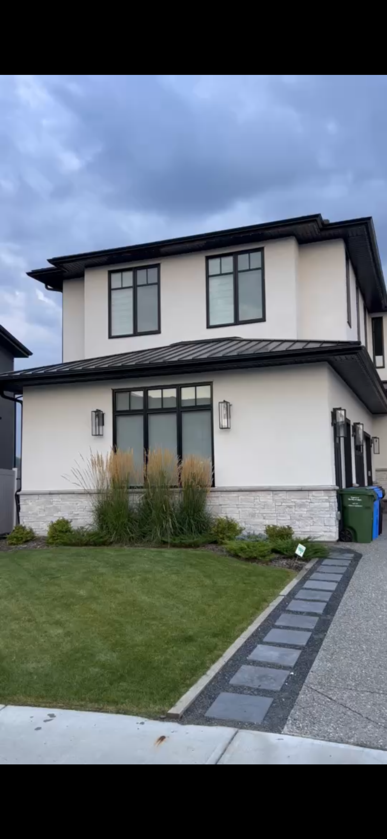 A modern two-story house with white walls, large black-framed windows, and a metal roof. There is a small front yard with green grass, ornamental grasses, and small shrubbery, and a stone pathway leading to the front door. Two outdoor wall-mounted light fixtures are on the front of the house. Trash bins are visible on the side.