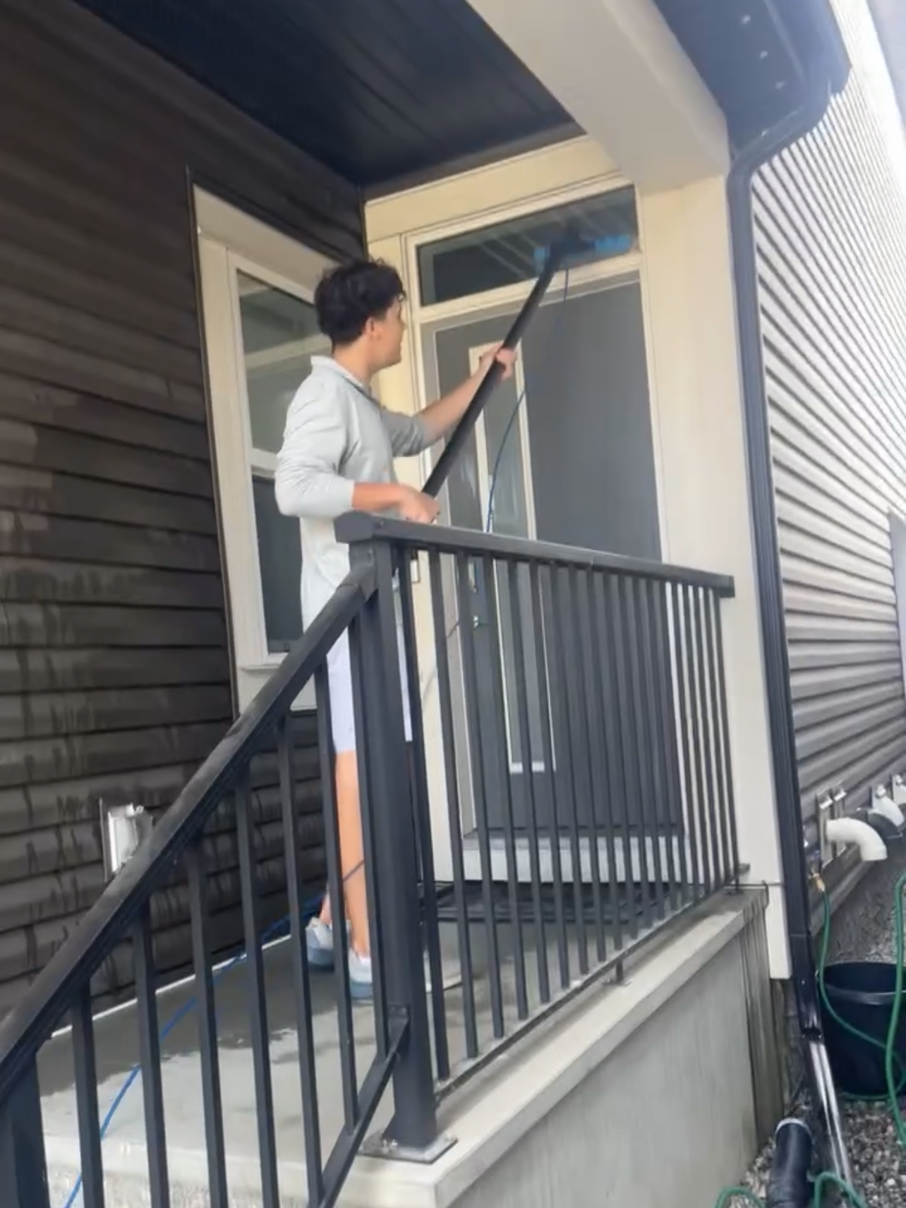 A woman cleaning the exterior window of a house's front door using a long-handled squeegee on a small porch with a black metal railing.