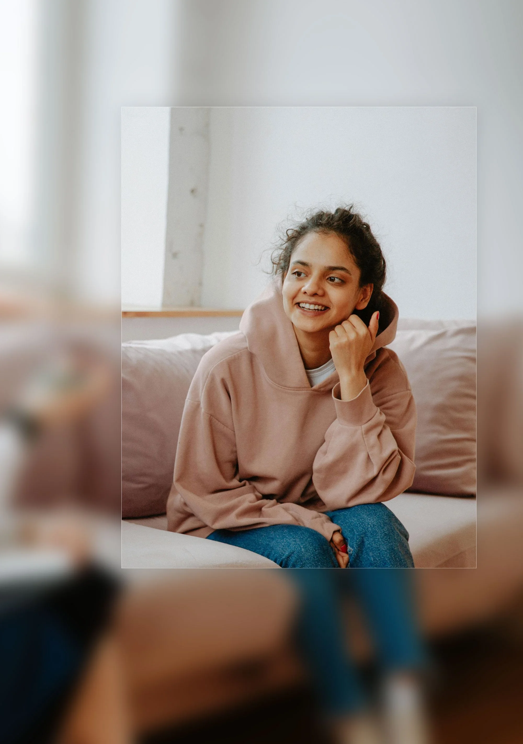 A young woman smiling, sitting on a beige sofa in a room with white walls. She is wearing a pink hoodie and blue jeans, with her hair tied back, speaking with a licensed-therapist.
