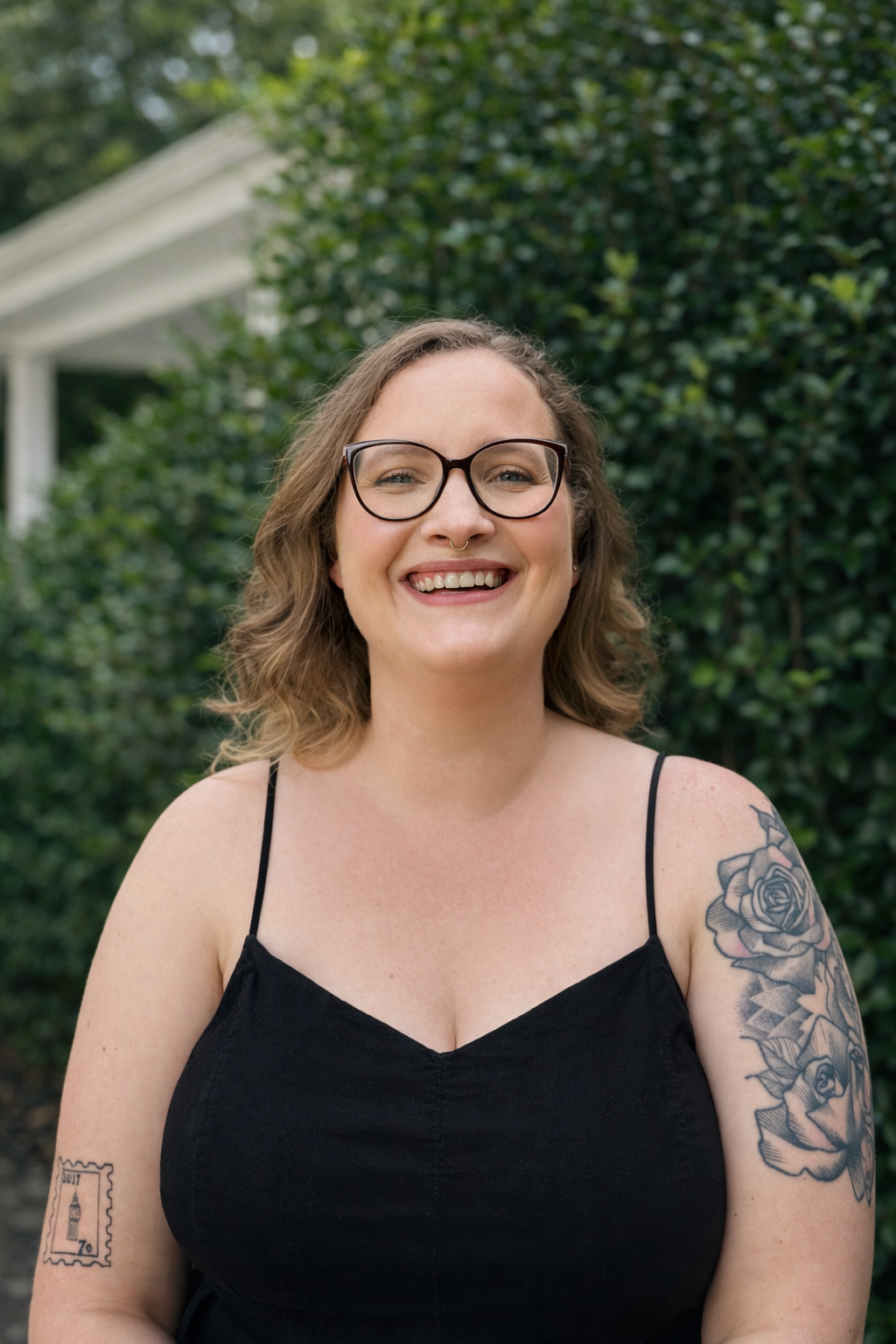 A smiling woman with glasses and tattoos outdoors, wearing a black spaghetti strap top.