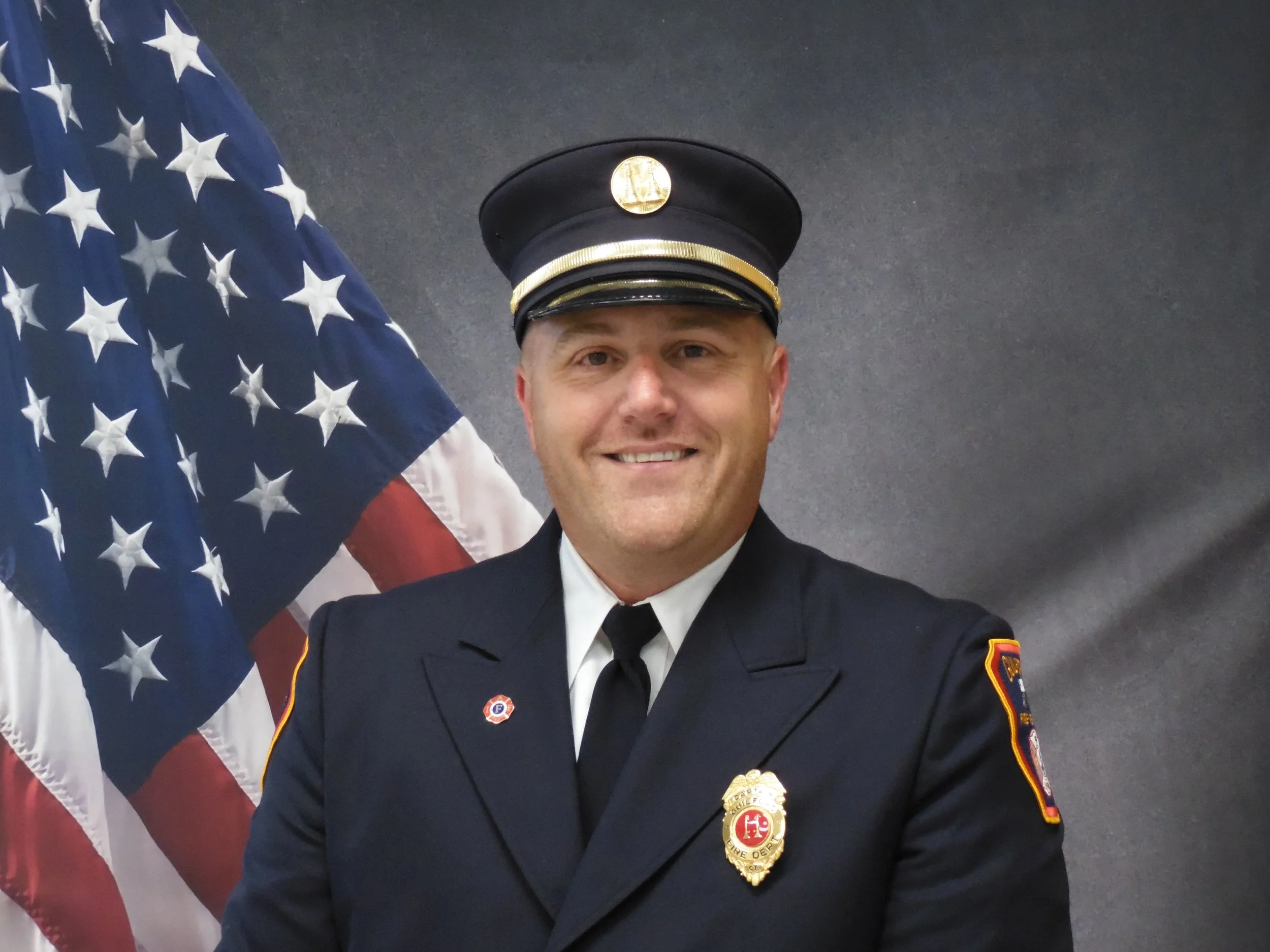 A smiling firefighter in uniform standing in front of an American flag. The firefighter is wearing a dark blue uniform with a badge and a cap with a gold emblem.