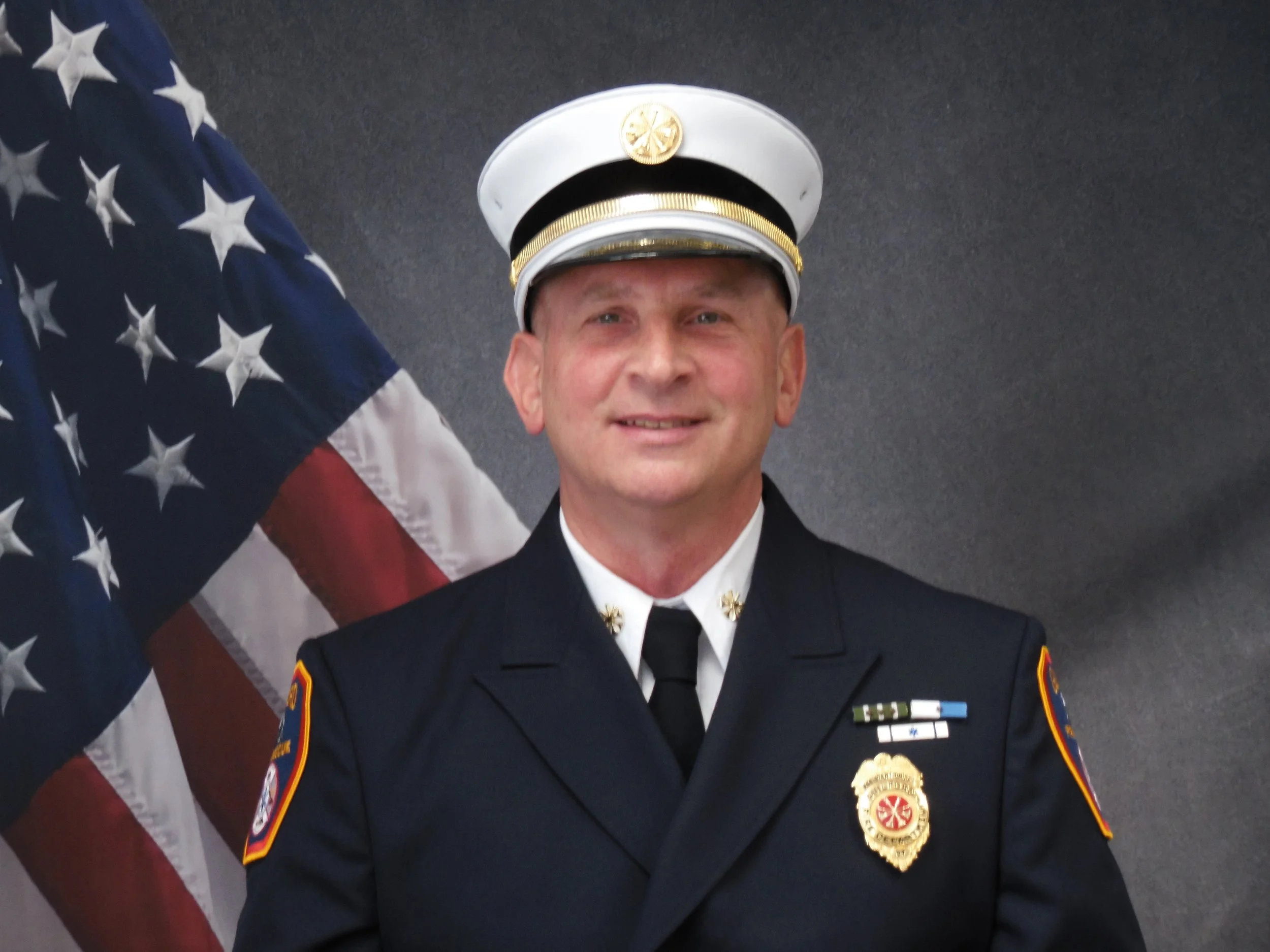 A man in a police uniform is smiling in front of a dark background, with partially visible American flag behind him.