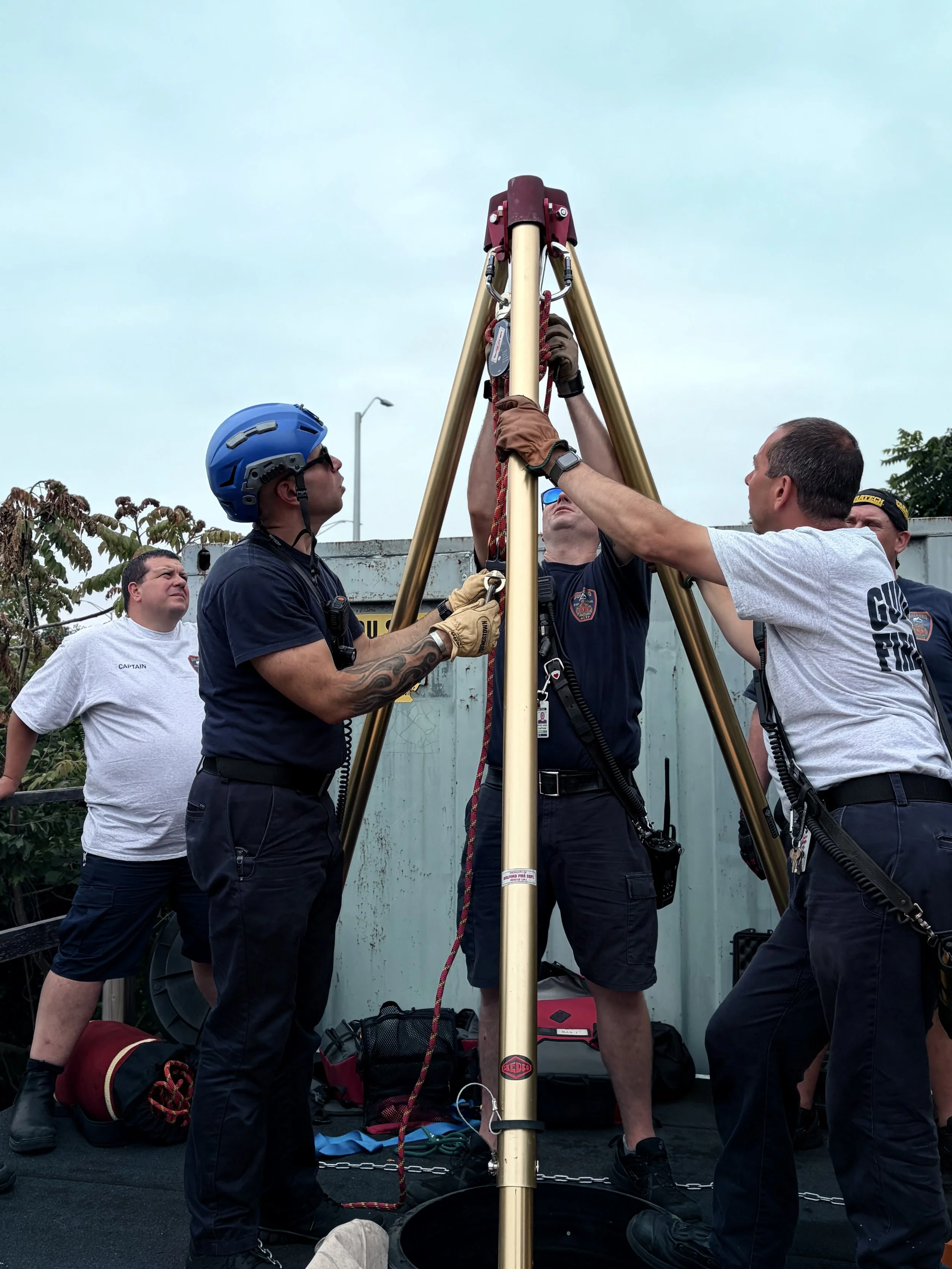 Group of people, some wearing firefighting uniforms, gathered outdoors, working on a tall tripod-like structure with a beige pole. They appear to be engaged in a rescue or training exercise.