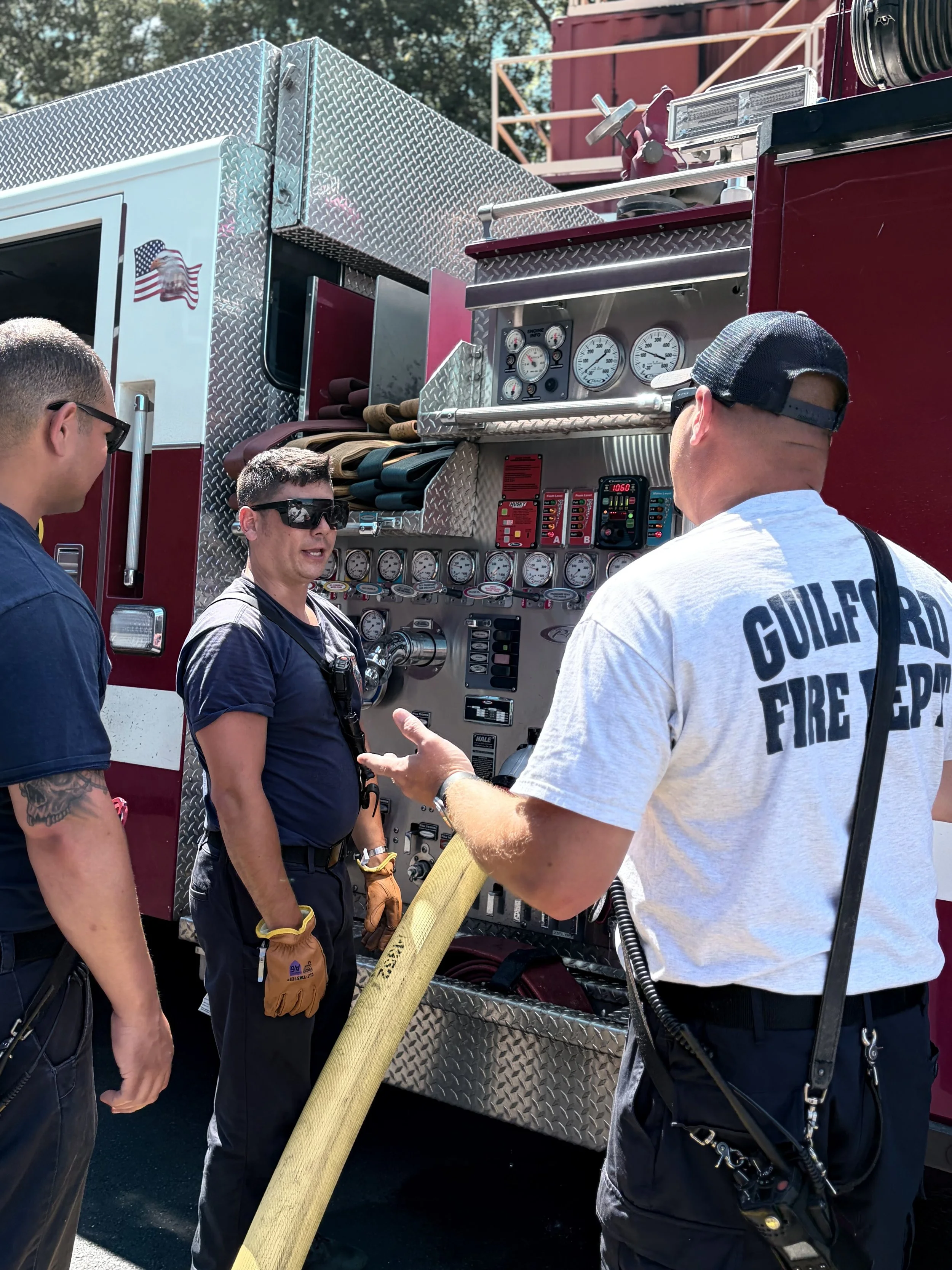 Firefighters and fire department personnel discuss and operate a fire truck with various gauges, hoses, and equipment during a demonstration or emergency call.