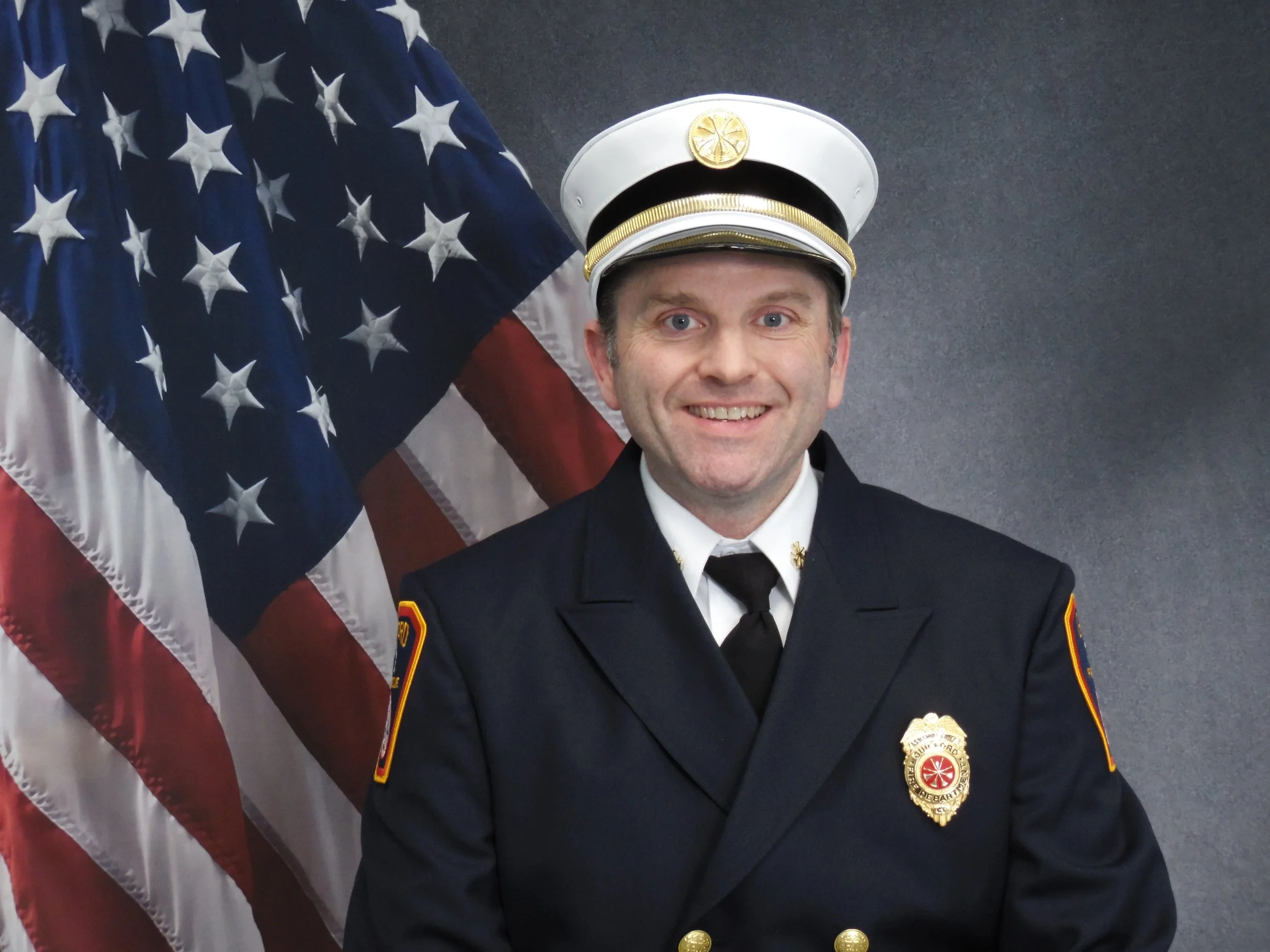 A smiling firefighter is standing in front of an American flag. He is wearing a formal firefighter uniform with a badge visible on his chest, and a white firefighter hat with a gold emblem.