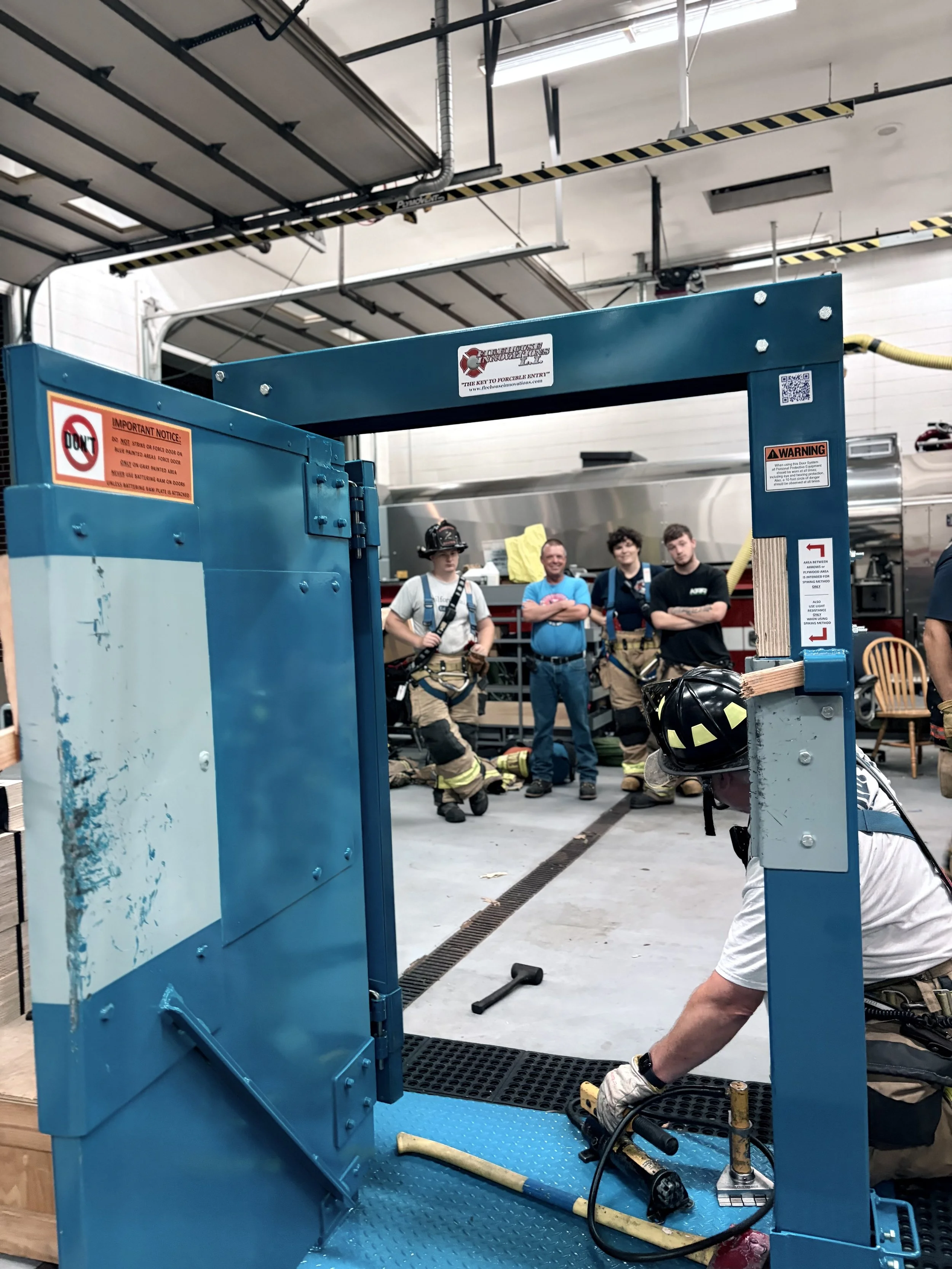 Firefighters and individuals in a garage working around a blue vehicle lift. Some are wearing firefighter gear, and some are in casual clothing. The garage has an open door and various equipment visible.